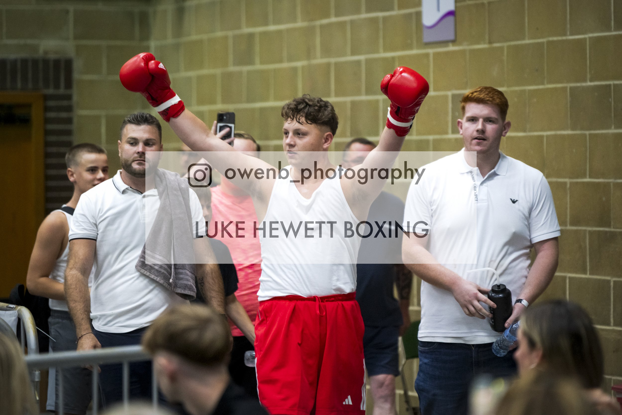 A young man in red boxing shorts and red gloves raises his arms in victory inside a gymnasium, surrounded by people, including a man in a white shirt and a young woman with a smartphone, with a brick wall in the background.
