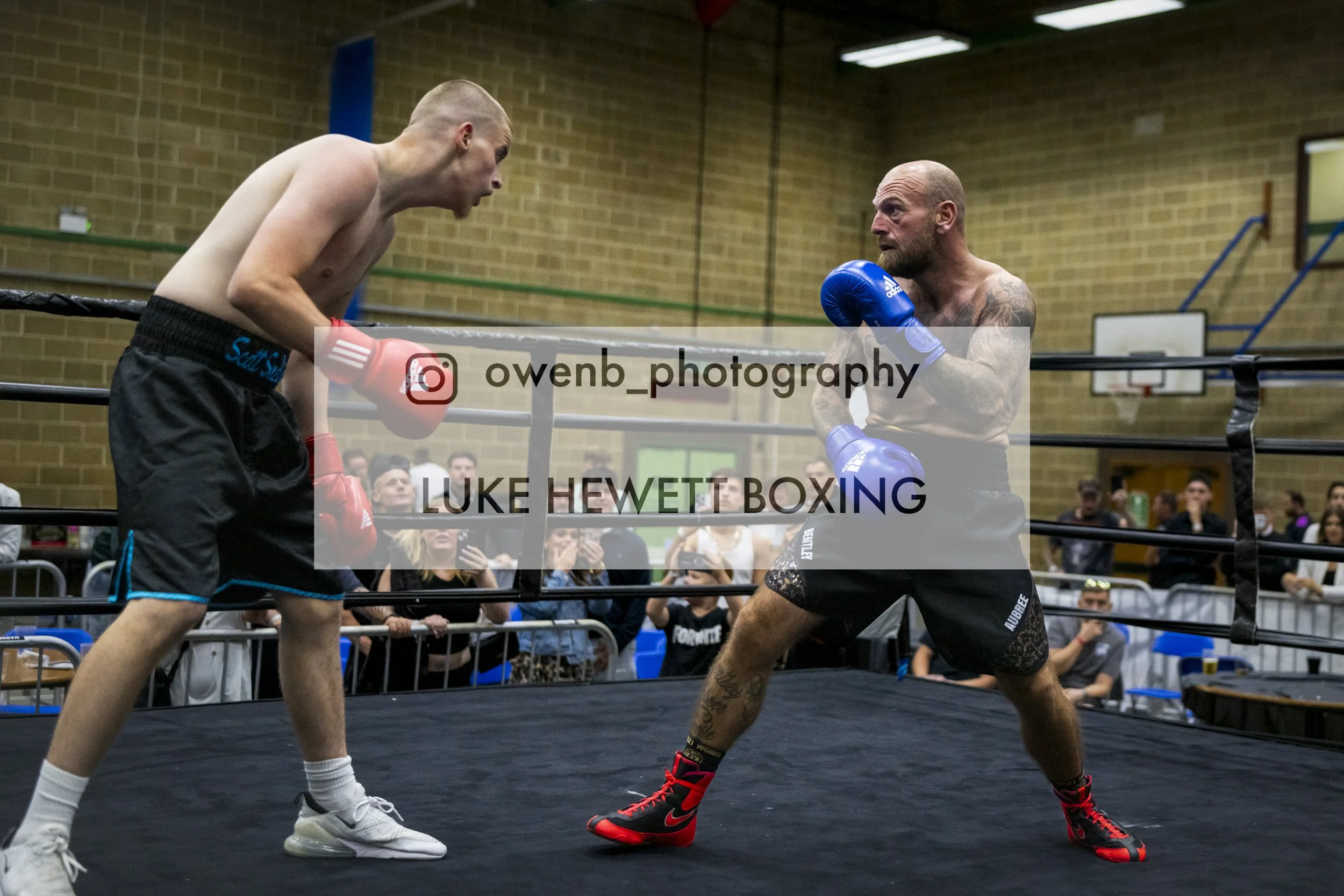 Two shirtless male boxers with gloves sparring in a boxing ring, with a crowd watching in the background. One appears older with tattoos; the other is younger, bald, and wearing black shorts with blue trim.