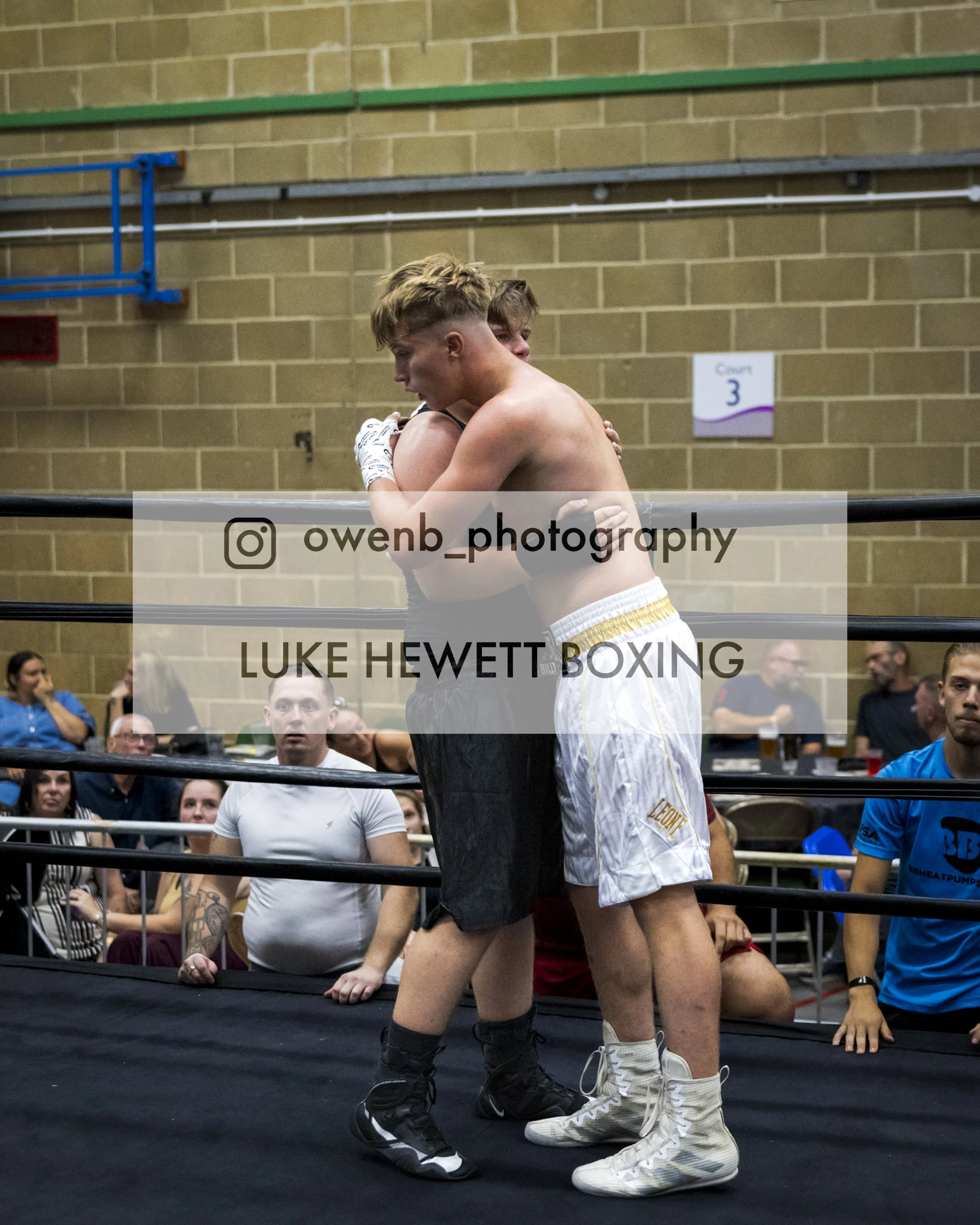 Two male boxers hugging inside a boxing ring during a match, with spectators watching from the background.