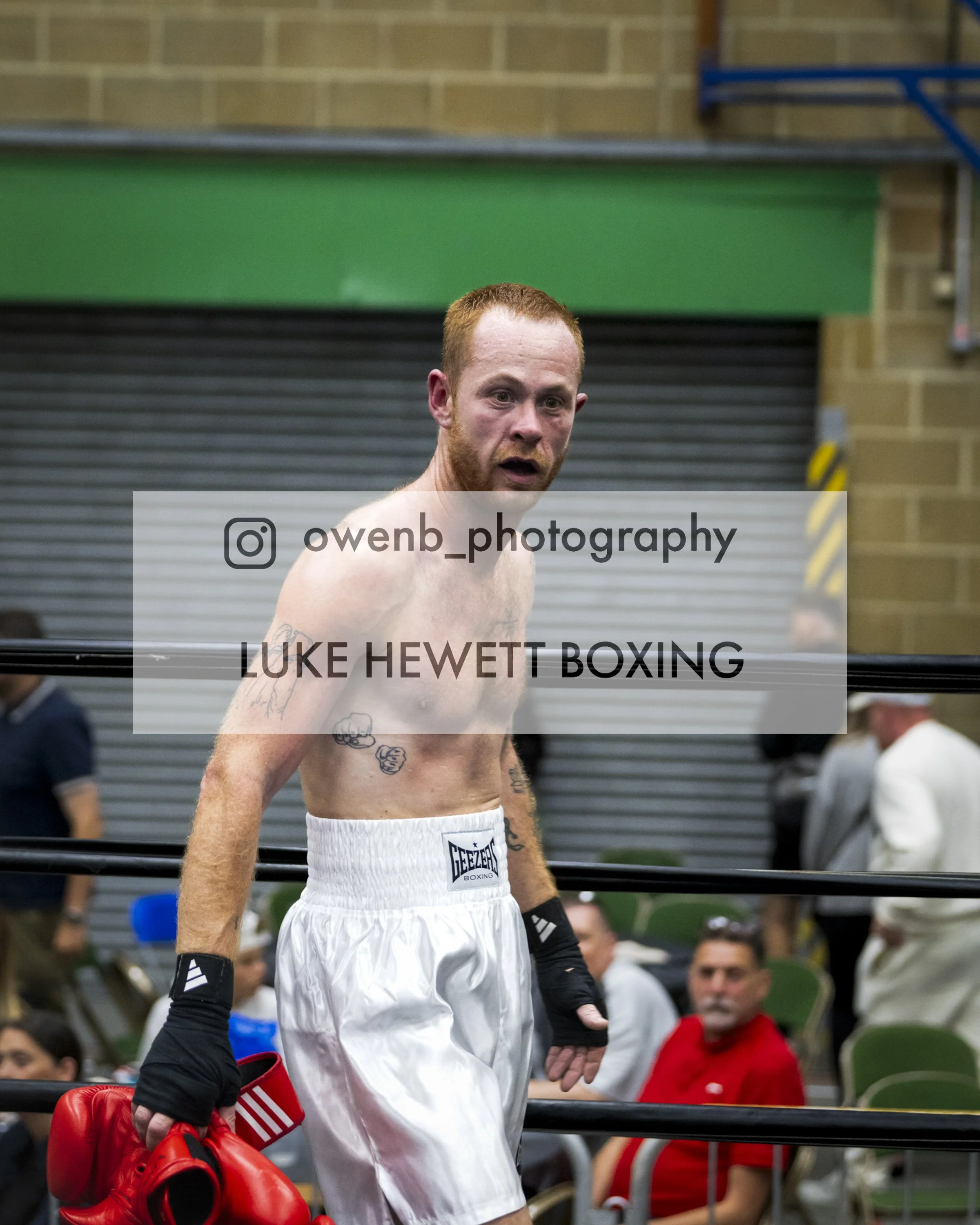 A shirtless male boxer with red hair and beard, wearing white boxing shorts and black arm sleeves, standing in a boxing ring with red boxing gloves hanging from his shoulder, in front of a blurred crowd.
