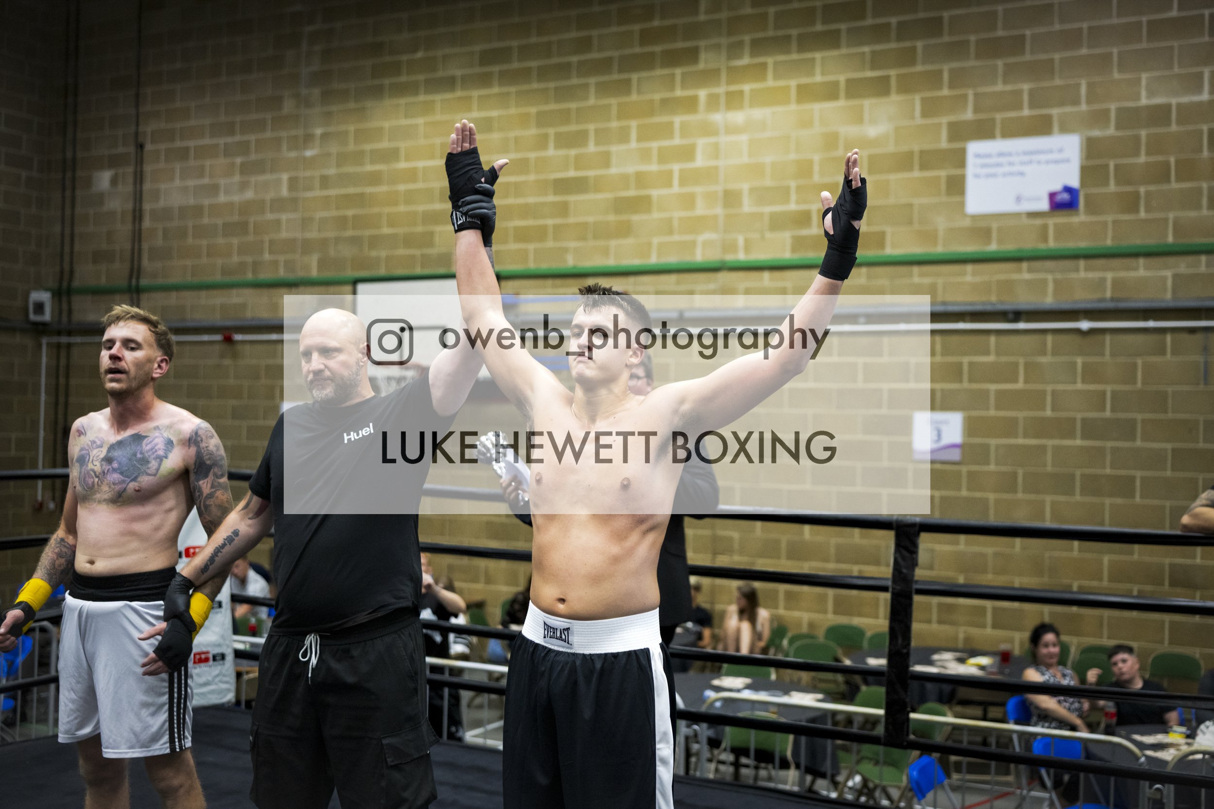 Boxer Luke Hewett with his arms raised in victory inside a boxing ring, surrounded by people and spectators.