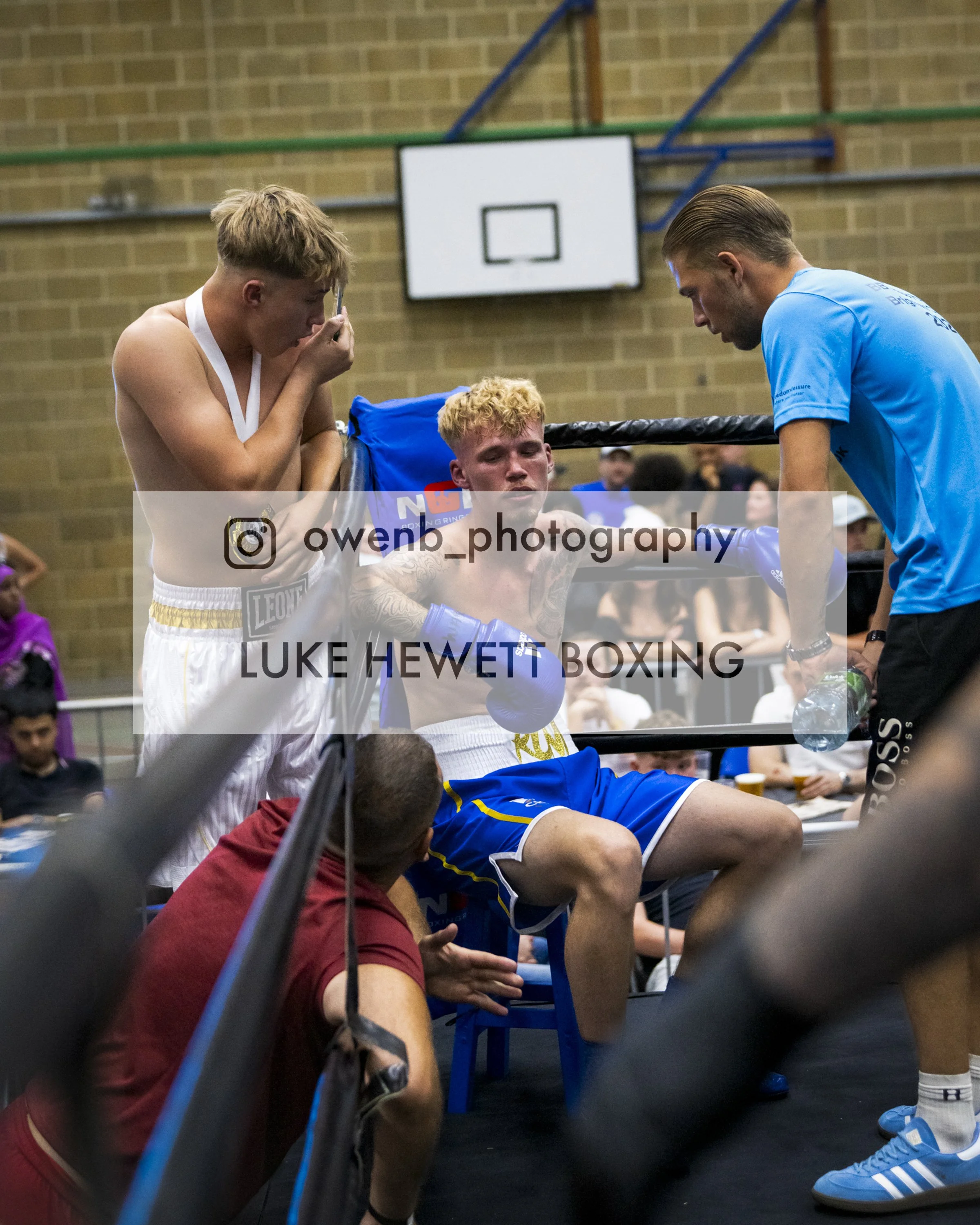 A group of young male boxers inside a boxing ring, with one seated and being attended to by a coach or trainer. The boxer is wearing blue shorts and blue boxing gloves. Another boxer in red shorts and a shirt is sitting at the edge of the ring, and t