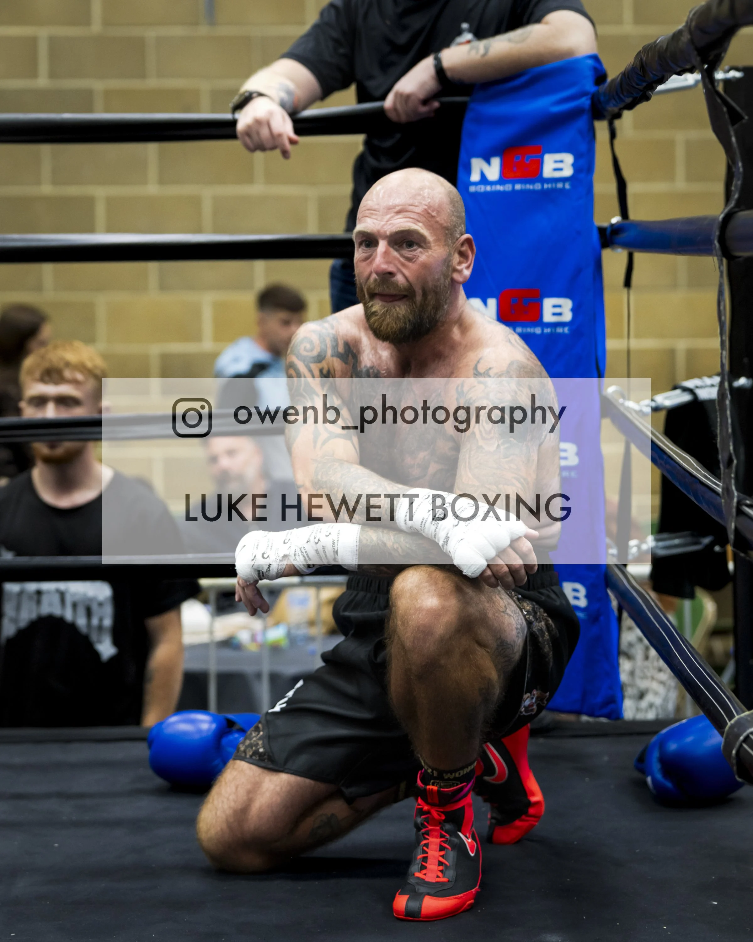A muscular, tattooed man with a beard training in a boxing gym, kneeling on the floor and wearing black shorts with red accents, black and red boxing shoes, protective hand wraps, and blue boxing gloves, with boxing ring ropes nearby.