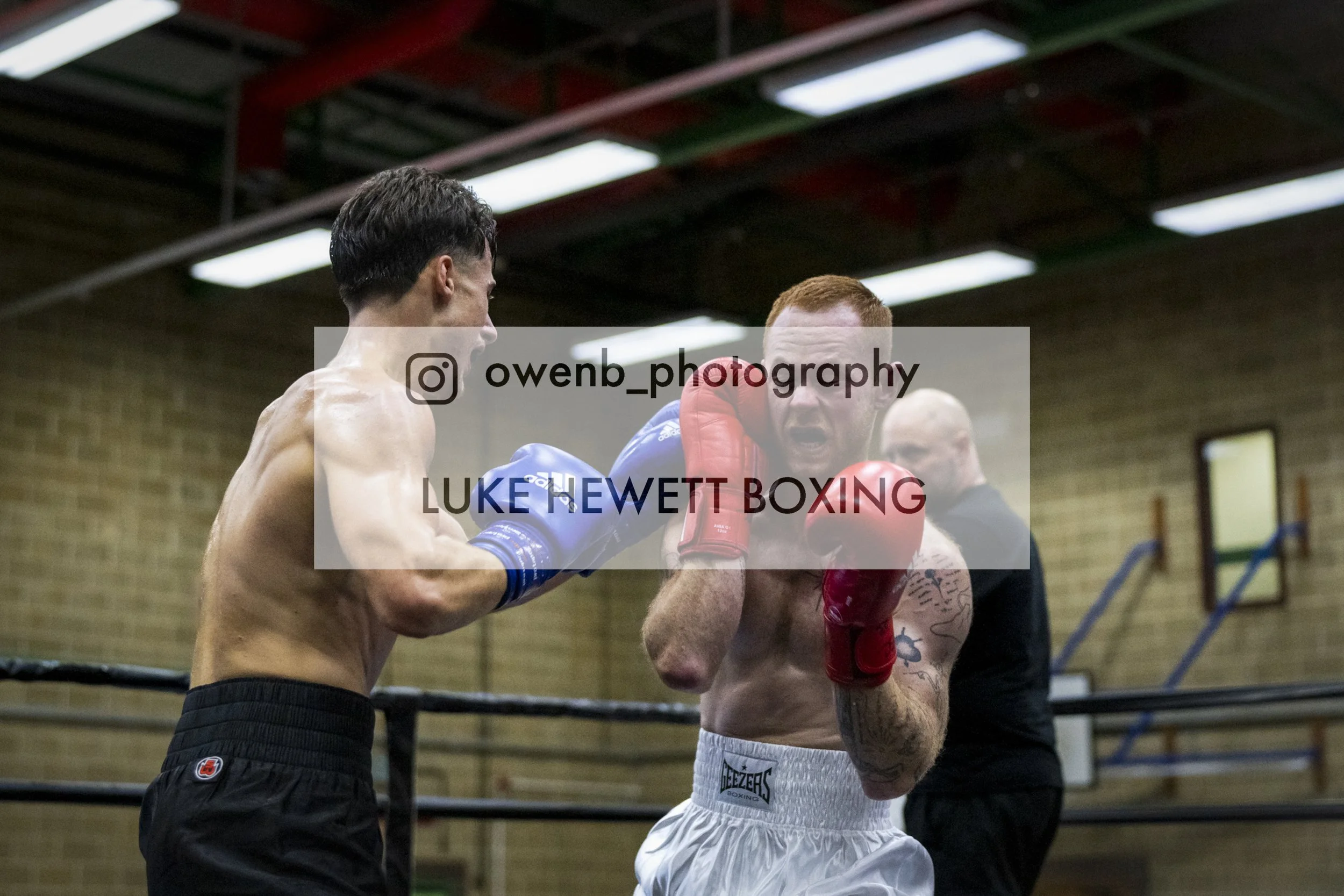 Two men training in a boxing gym. One man is shirtless with boxing gloves, punching a target held by the other man who is wearing a blue shirt and red gloves. The training session takes place in a gym with brick walls and wooden ceiling.