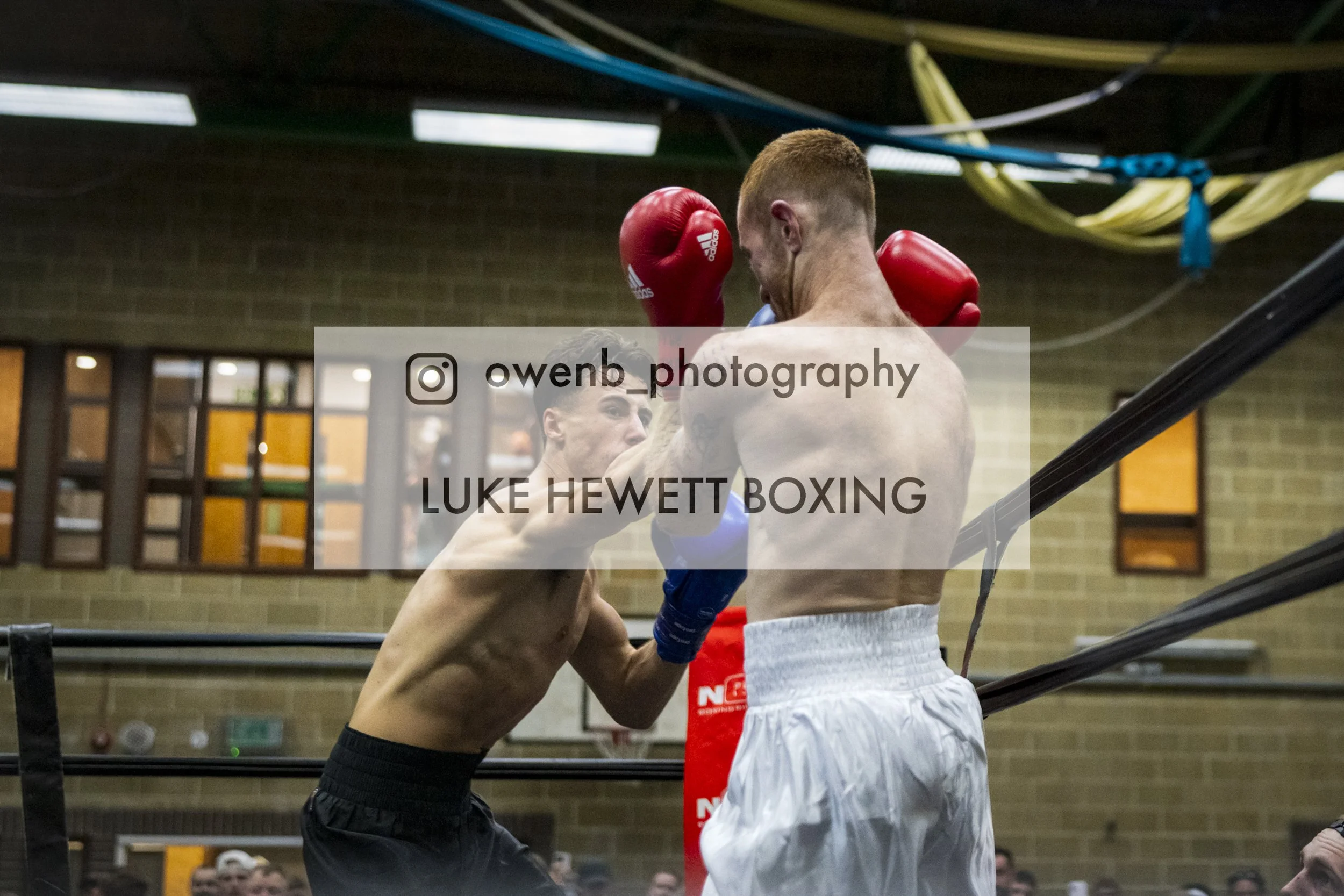 Two male boxers are engaged in a boxing match inside a ring. One is wearing red gloves and white shorts, while the other is wearing black shorts. The boxer with red gloves is leaning forward, with his forehead touching the forehead of the other boxer