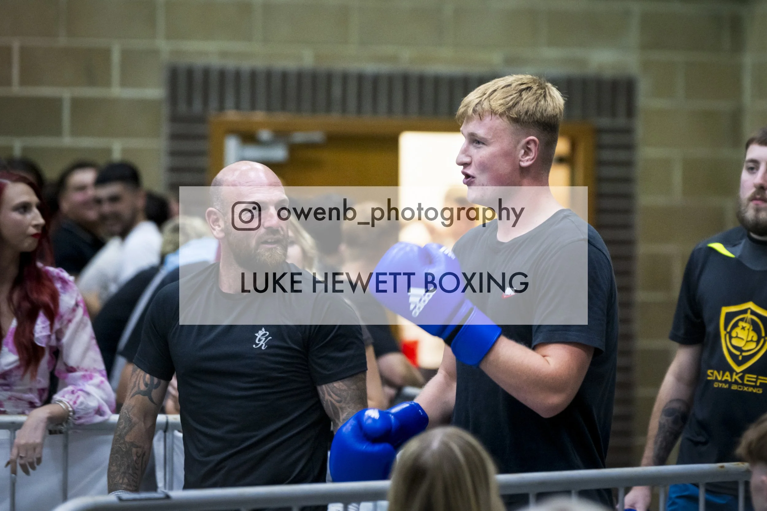 Boxer Luke Hewett in a gym with fans and trainers, wearing blue boxing gloves, engaged in a sparring session or training.