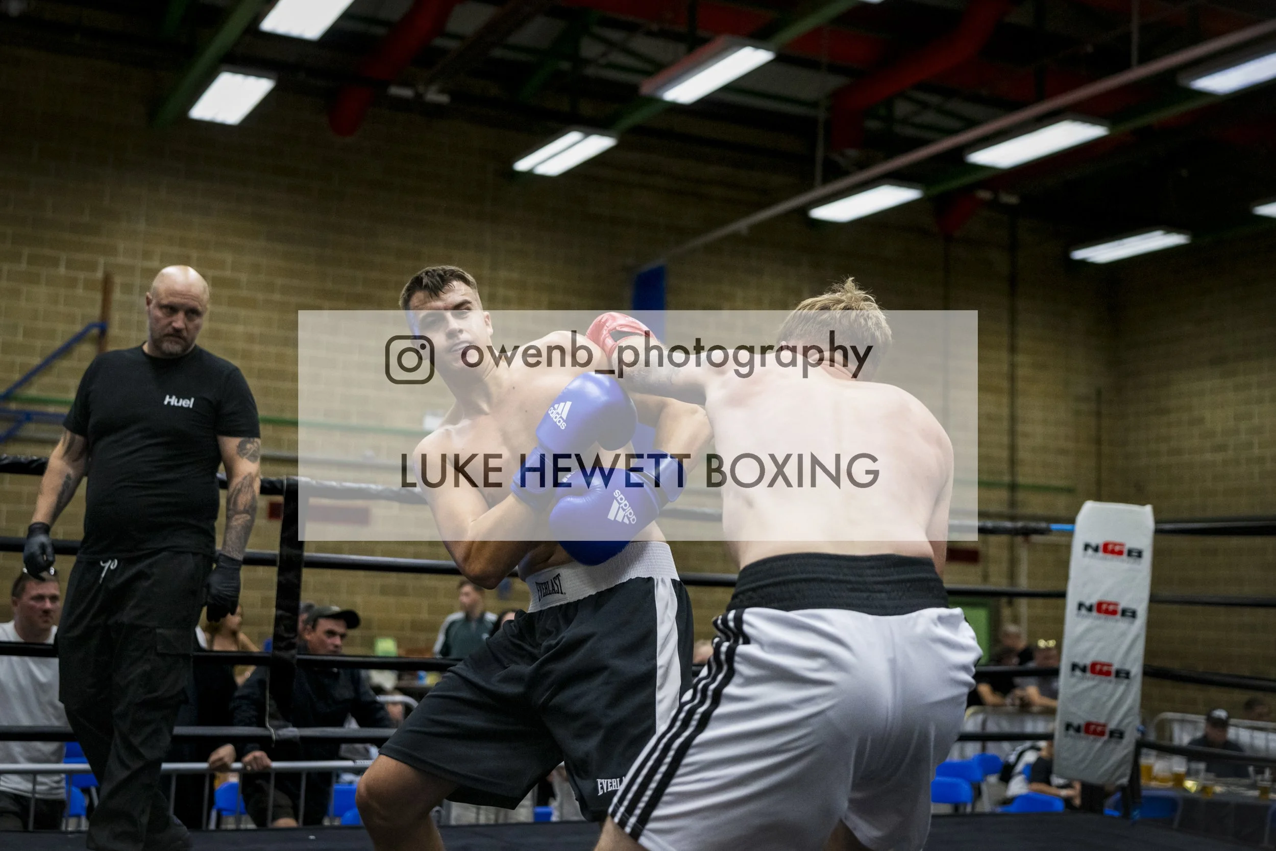 Two male boxers are engaged in a match inside a boxing ring, with one delivering a punch to the other's face. A referee stands nearby observing the fight, while spectators watch from the background.