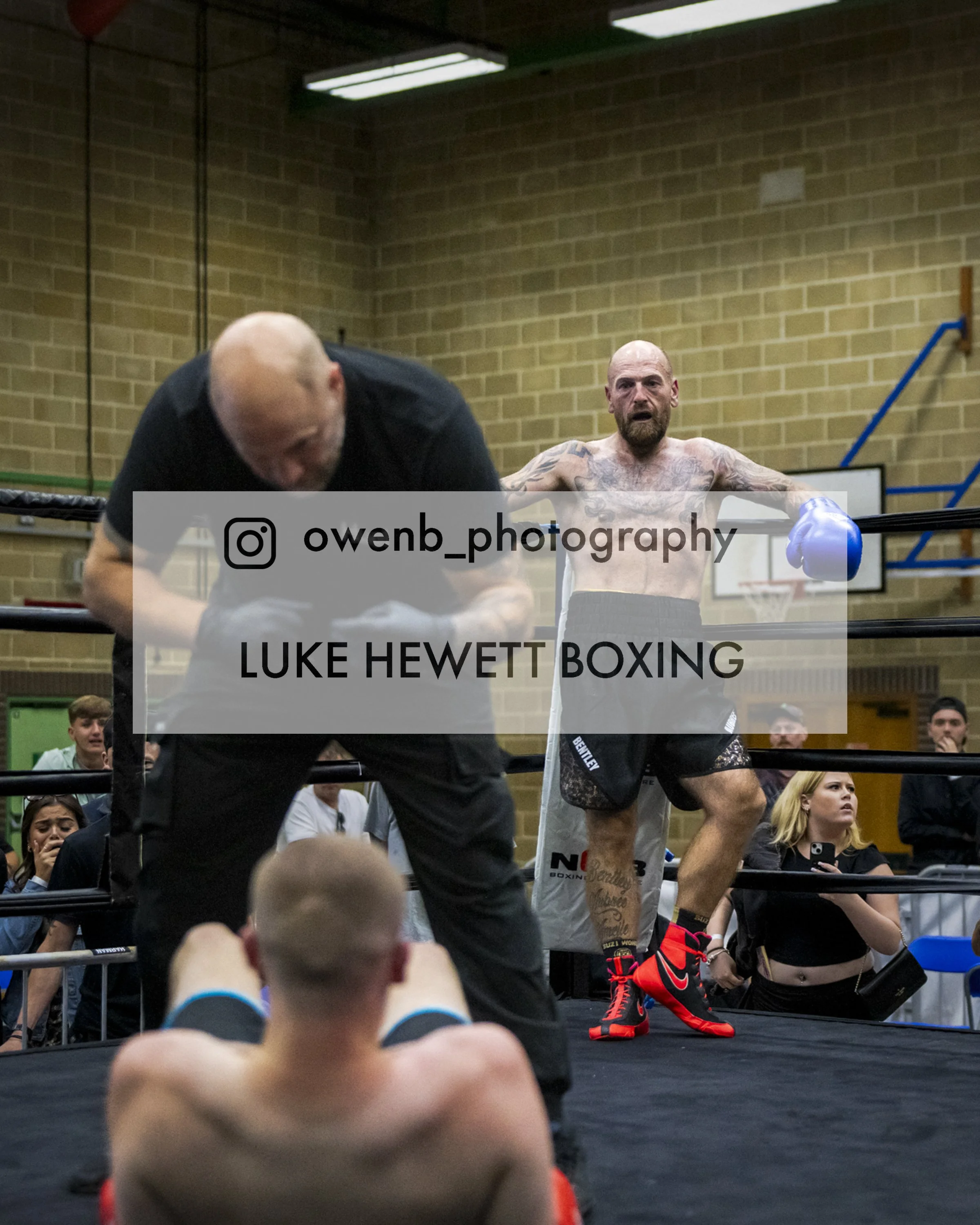 A boxer in boxing shorts and gloves standing in a boxing ring with his arms out, while a trainer leans over in front of him. Spectators seated around the ring watch the scene.