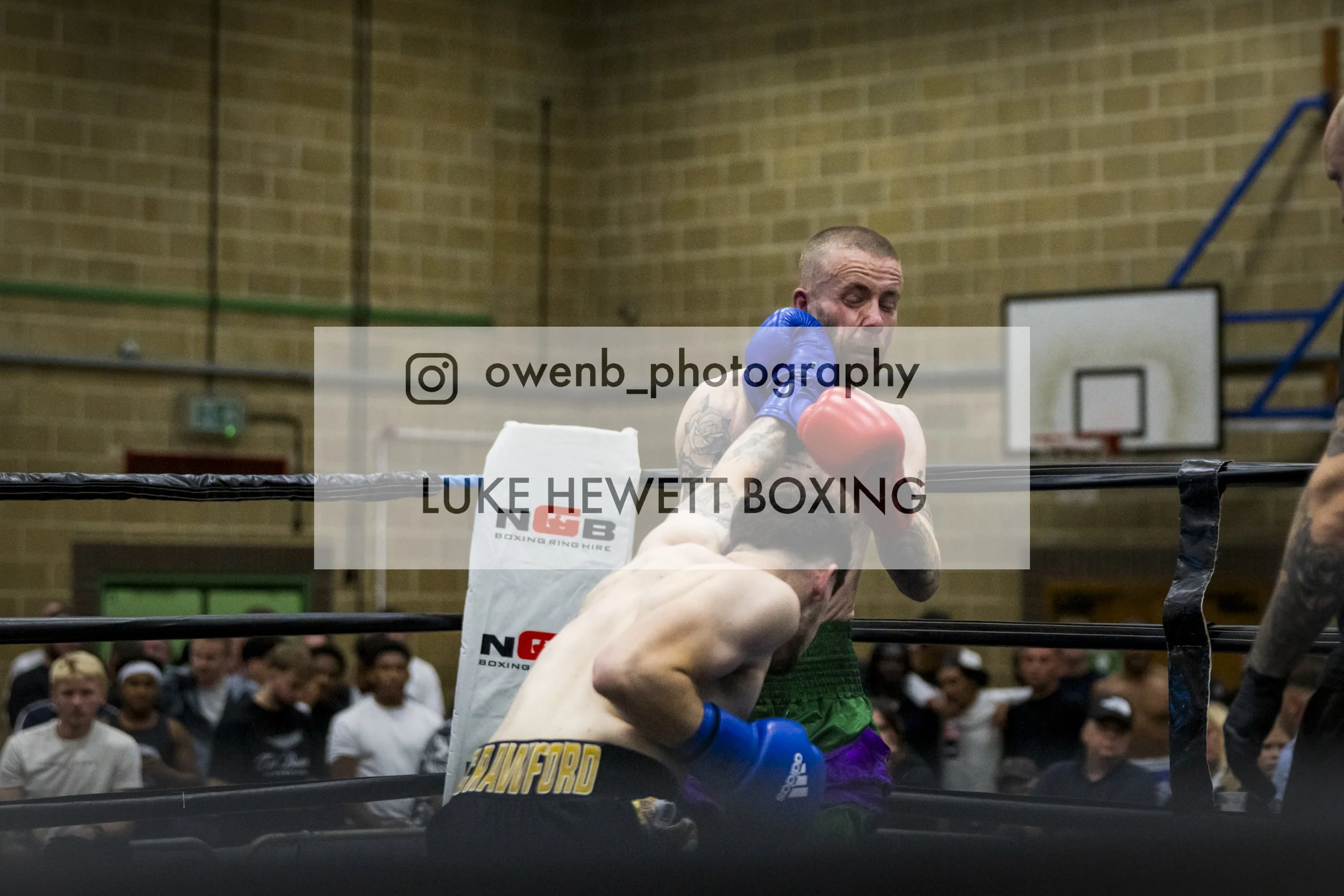 Two boxers in a ring, one with blue gloves and the other with red gloves, engaged in a match. The boxer with red gloves is leaning against the ropes, and the scene is set in a gymnasium with a brick wall and an audience in the background.