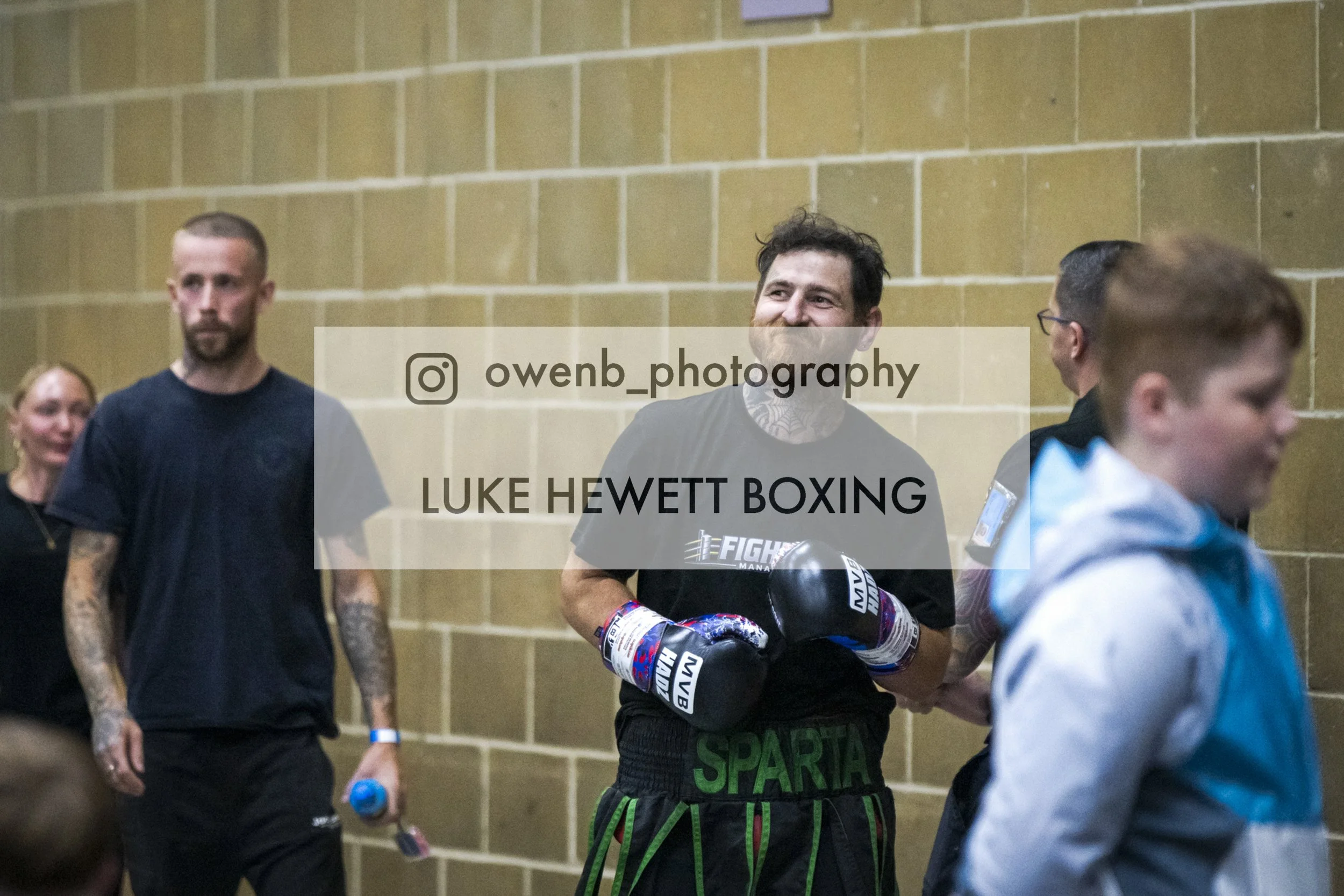A group of people in a gym or training facility, with a man wearing boxing gloves in the foreground, smiling. Others are standing nearby, some holding water bottles. The background features a beige brick wall. The text overlay reads: "@owenb_photogra