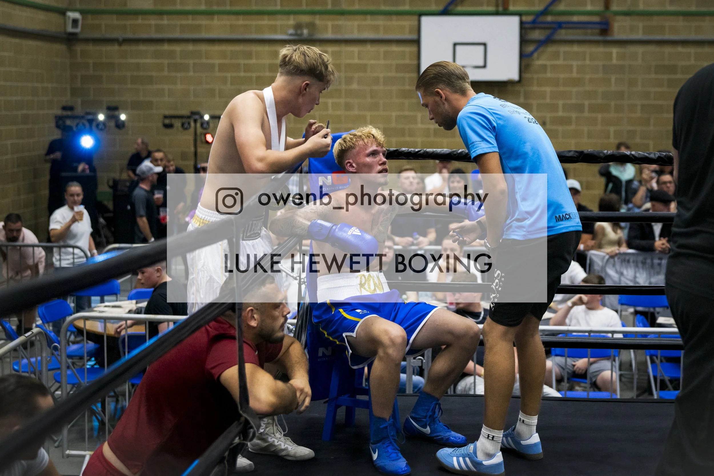 Boxer sitting in corner of boxing ring with coach and two other men, surrounded by spectators in an indoor gym with brick wall and basketball hoop in background.