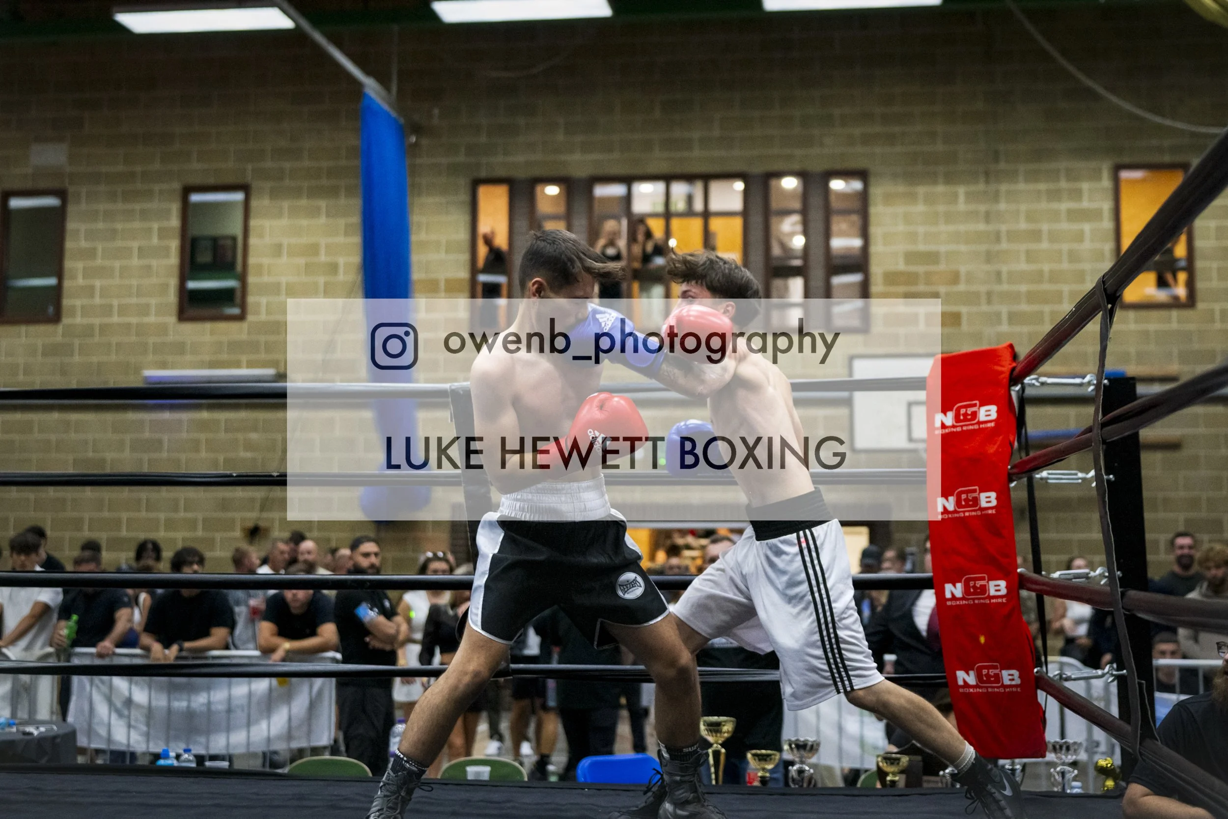 Two male boxers engaged in a match inside a boxing ring, surrounded by spectators and officials, with one boxer throwing a punch at the other.