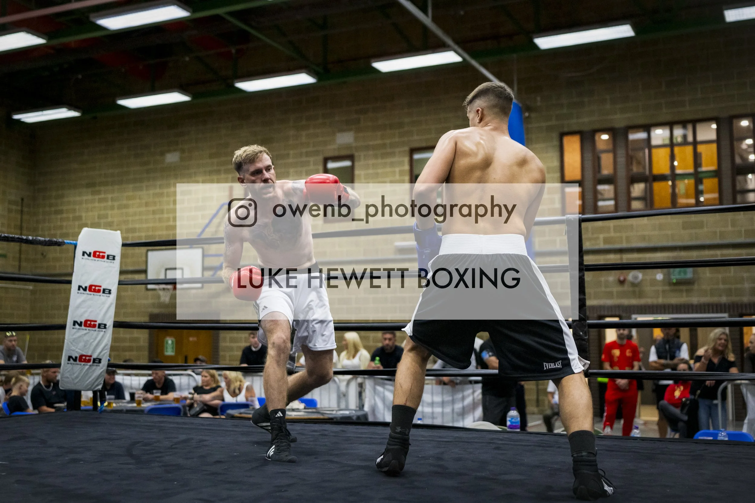 Two male boxers are engaged in a boxing match inside a ring in a gymnasium. One boxer is throwing a punch while the other is facing him with his back to the camera. Spectators are seated around the ring.