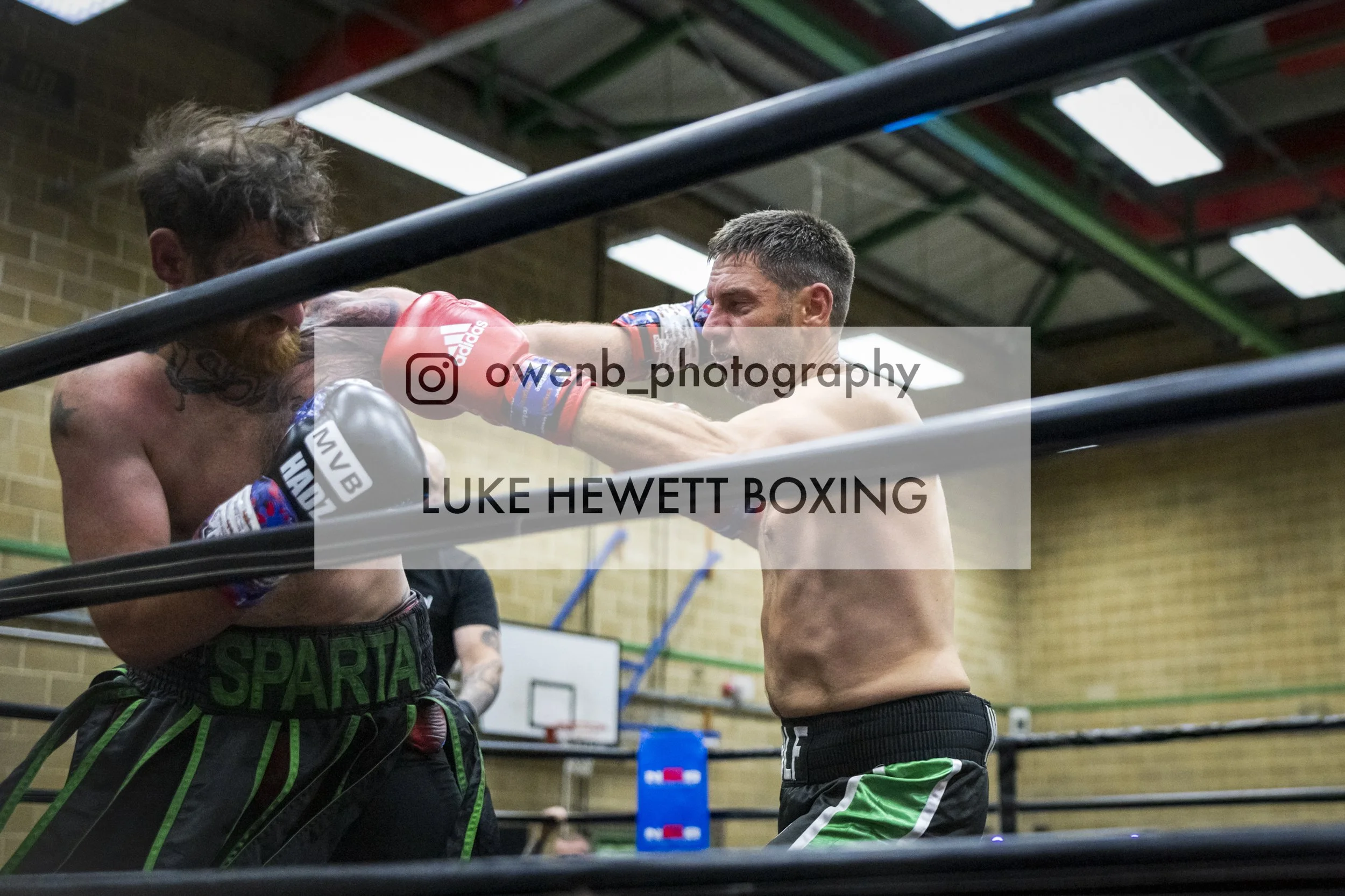 Two male boxers are sparring in a boxing ring, with one throwing a punch. Both are shirtless and wearing boxing gloves. The scene is inside a gymnasium with a basketball hoop visible in the background.