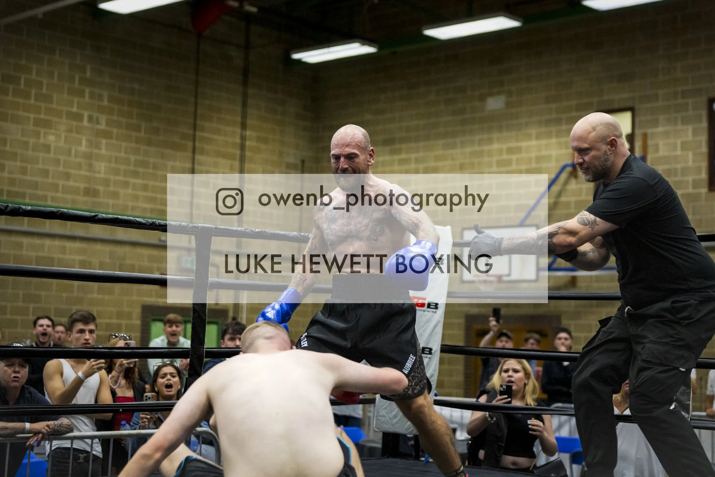 Boxing match inside a gym with an audience watching. A shirtless boxer with gloves is on the ground, while another boxer is standing nearby. The crowd shows various reactions, some excited and others surprised.