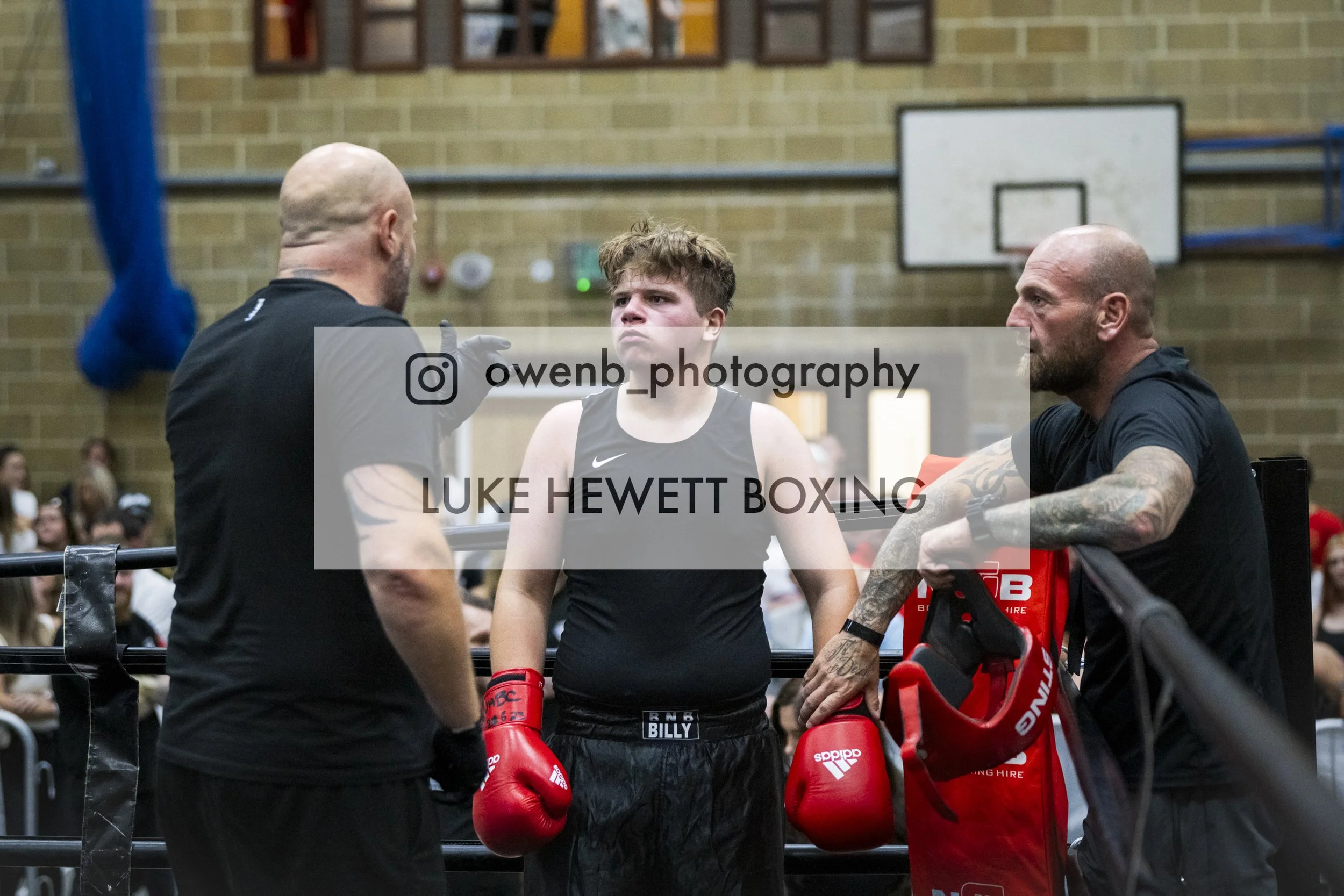 A young male boxer with red gloves and black shorts stands in a boxing ring, being advised by two adult men, one with a tattooed arm, during a boxing event in a gymnasium with a crowd in the background.