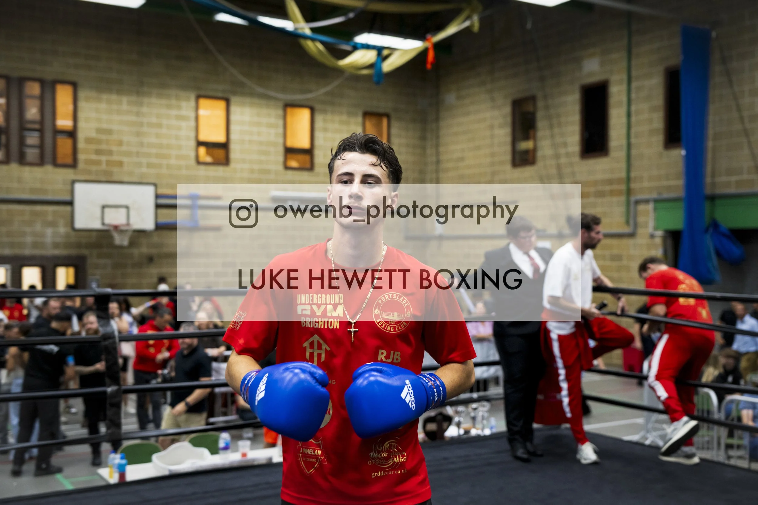 A young male boxer wearing blue gloves, a red shirt with logos, and a cross necklace, stands inside a boxing ring at a gym or sports hall. There are other people in the background, some in boxing or sports attire, and a basketball hoop can be seen on