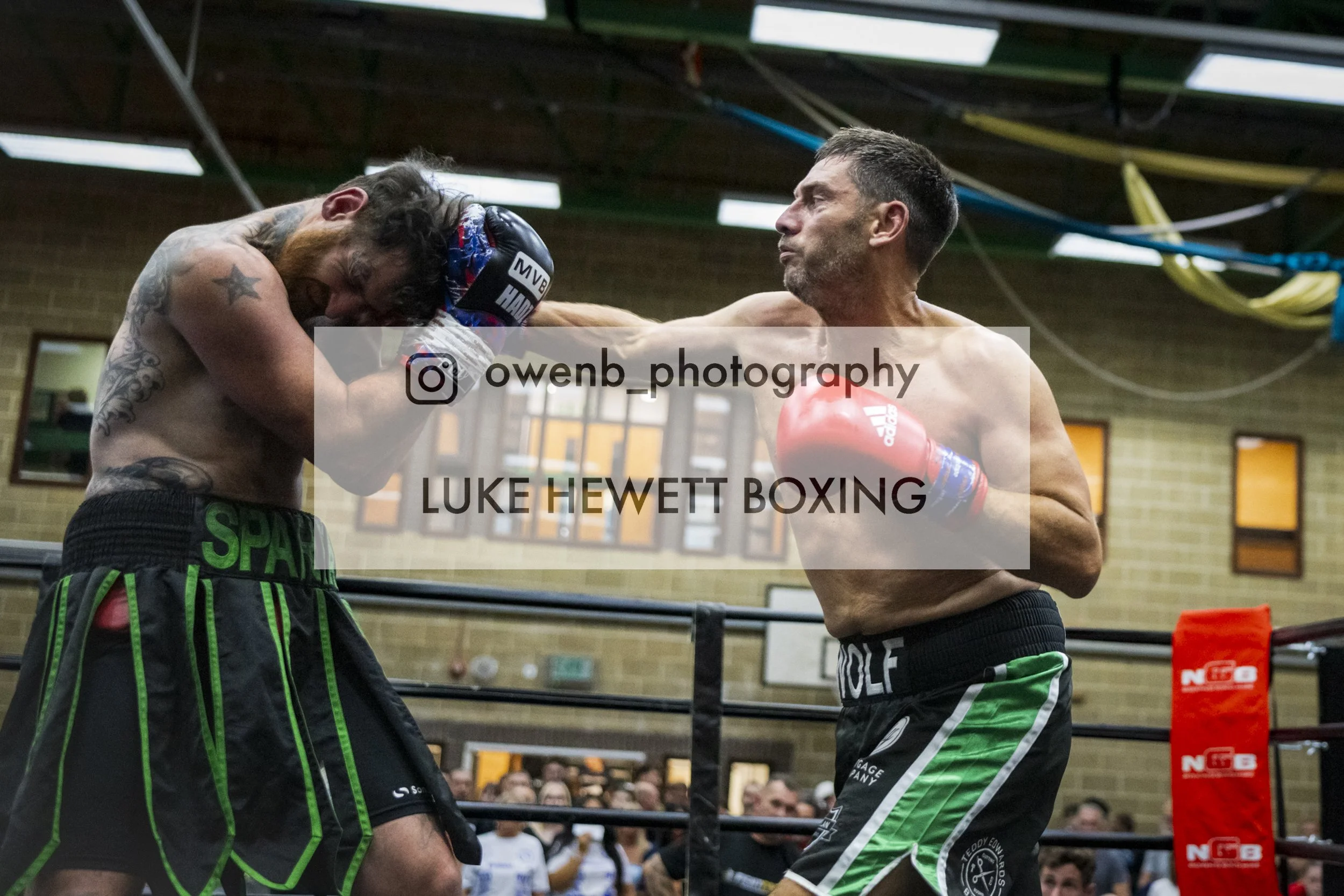 Two shirtless men boxing in a gym, one throwing a punch at the other, with spectators watching in the background.