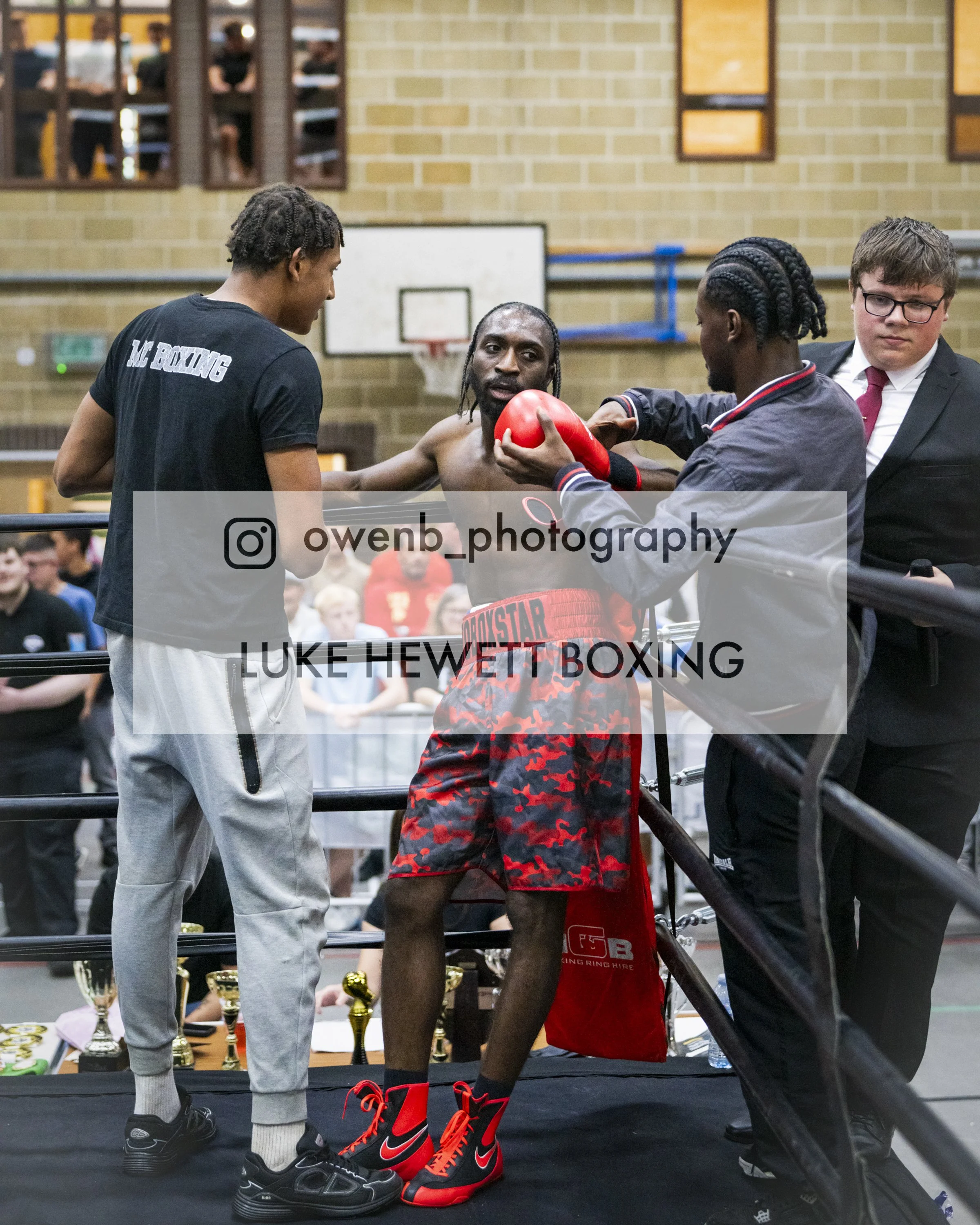 Boxer or kickboxer standing in the ring with two men, one handing him a red boxing glove, and the other presenting trophies in the background. The boxer is wearing red boxing shorts and red boxing shoes.