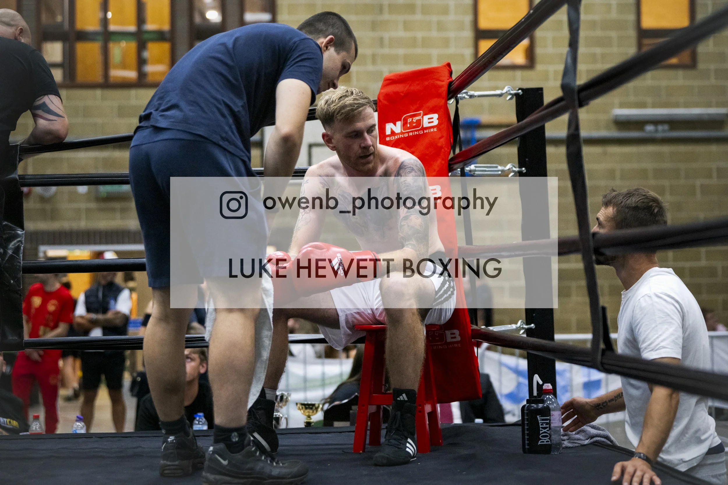 A boxing match scene where a trainer is attending to a seated, shirtless boxer with tattoos, who appears to be exhausted. The scene is inside a boxing ring with a red corner pad that has the logo 'NGB Boxing Ring Hire'. Other individuals are visible 