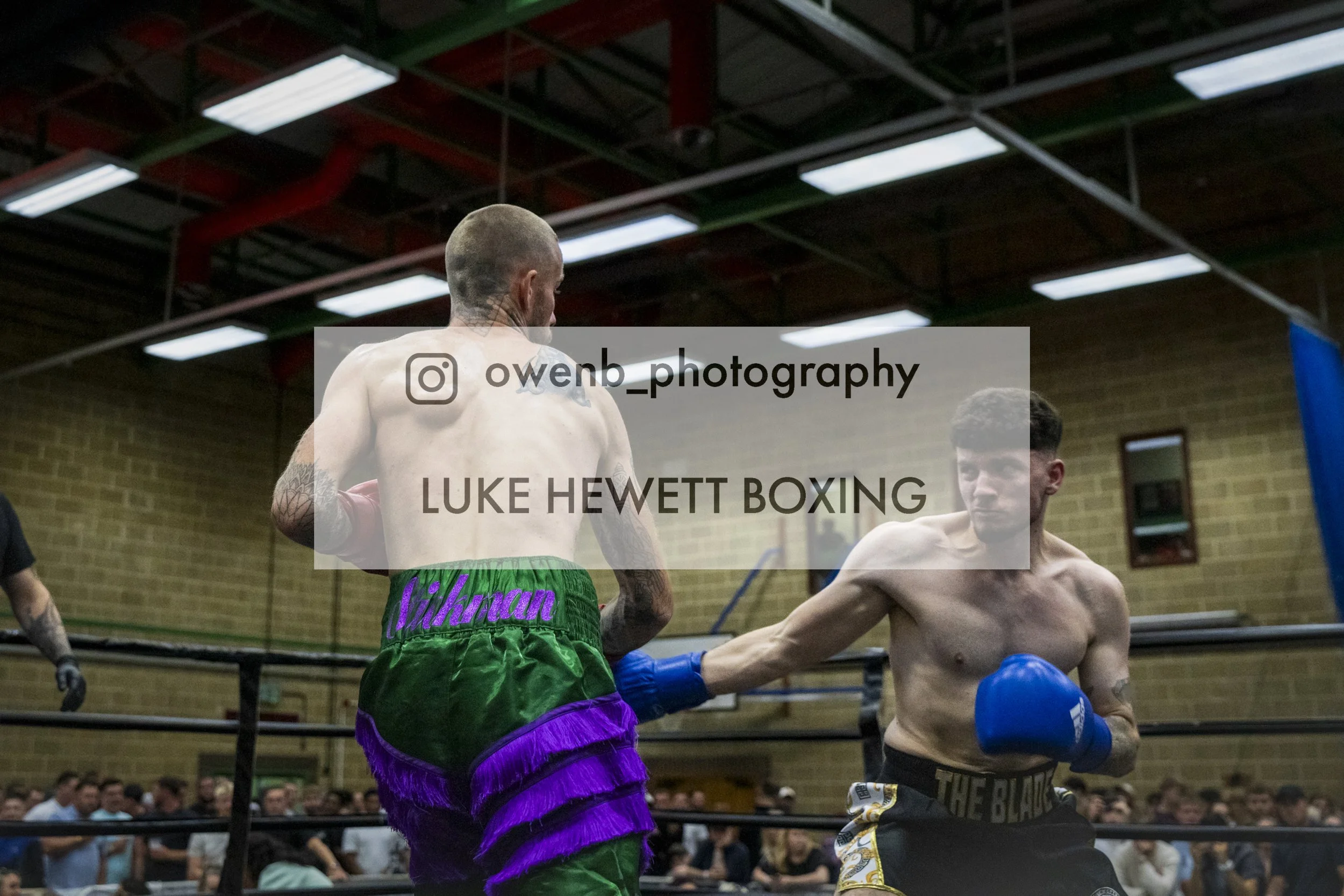 Two male boxers in a boxing ring, one in green and purple shorts and the other in black shorts, engaged in a match with an audience watching in the background.