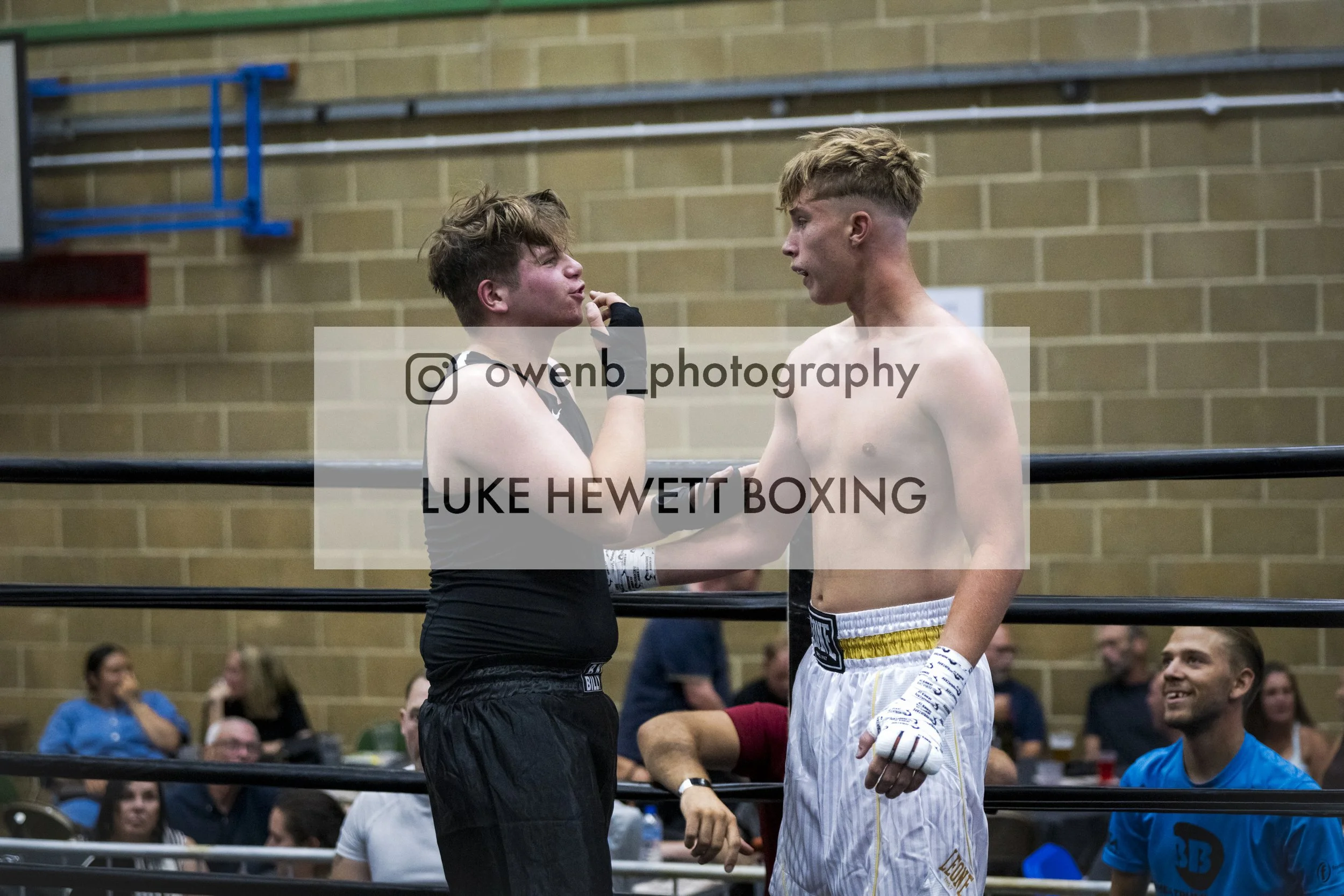 Two shirtless young male boxers facing each other inside a boxing ring, one in black shorts with a glove on his cheek, gesturing with his finger, and the other in white shorts with yellow accents, listening. Spectators in the background watch.