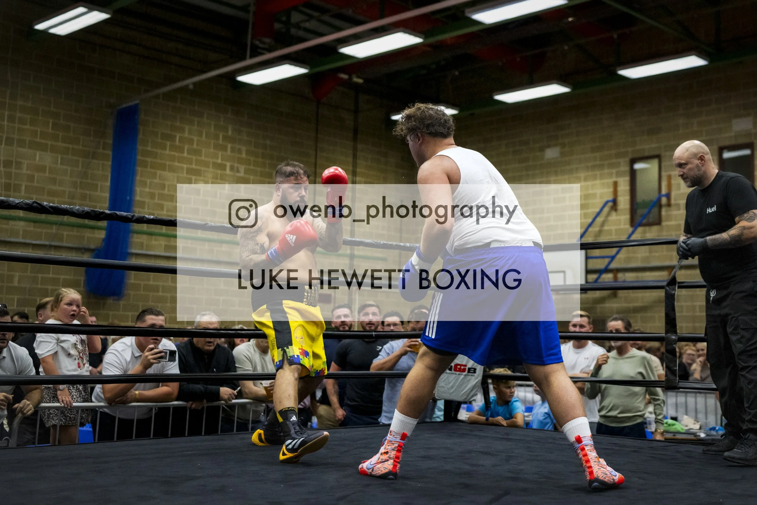 Two male boxers in a ring, with one in colorful shorts and the other in a white tank top and blue shorts, practicing or sparring while a referee stands nearby. Audience members watch from outside the ring, some recording or taking pictures.