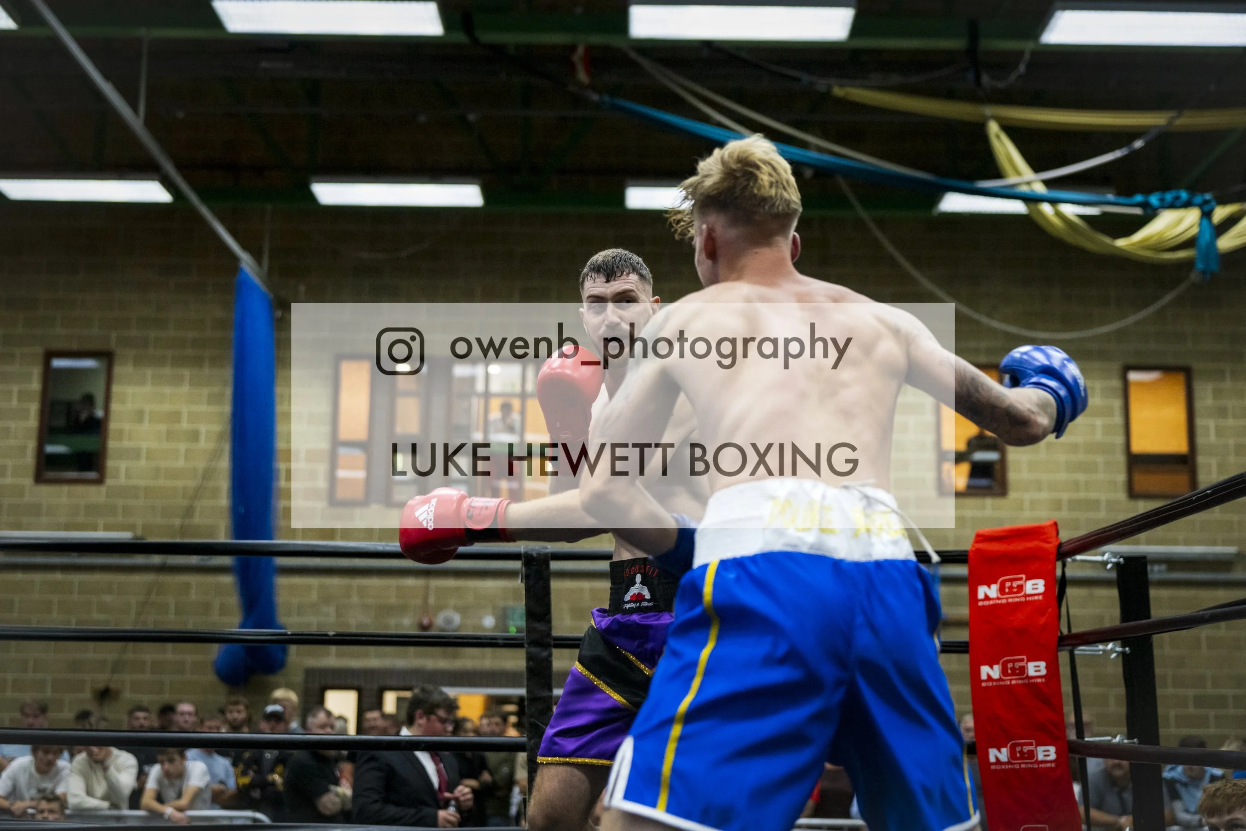 Two shirtless male boxers engaged in a boxing match inside a boxing ring with a crowd of spectators watching.