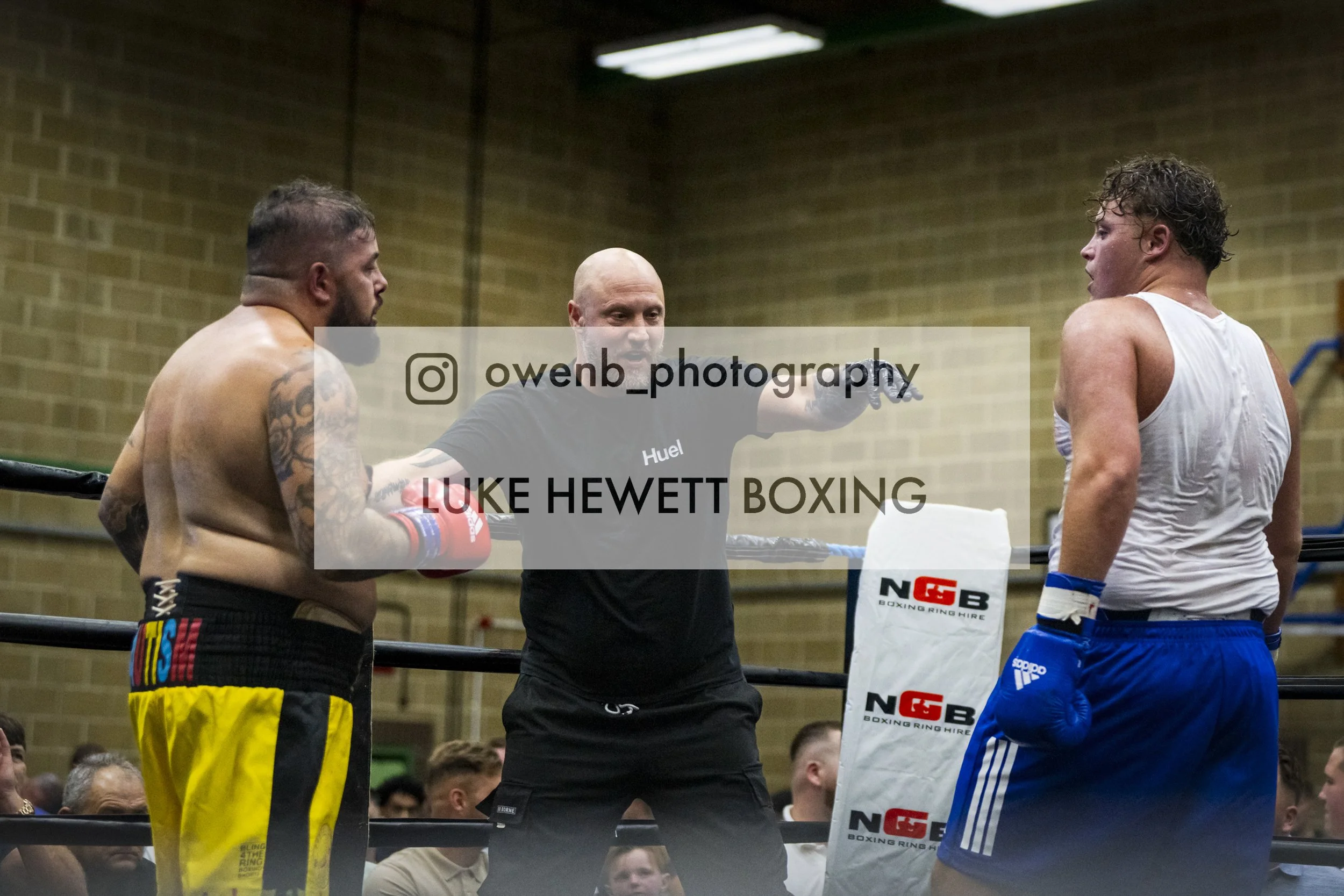 A boxing match in progress with two fighters in the ring and a referee in the middle, overseeing the fighters' exchange. The fighter on the left has tattoos and wears yellow trunks, while the fighter on the right has curly hair and wears blue trunks.