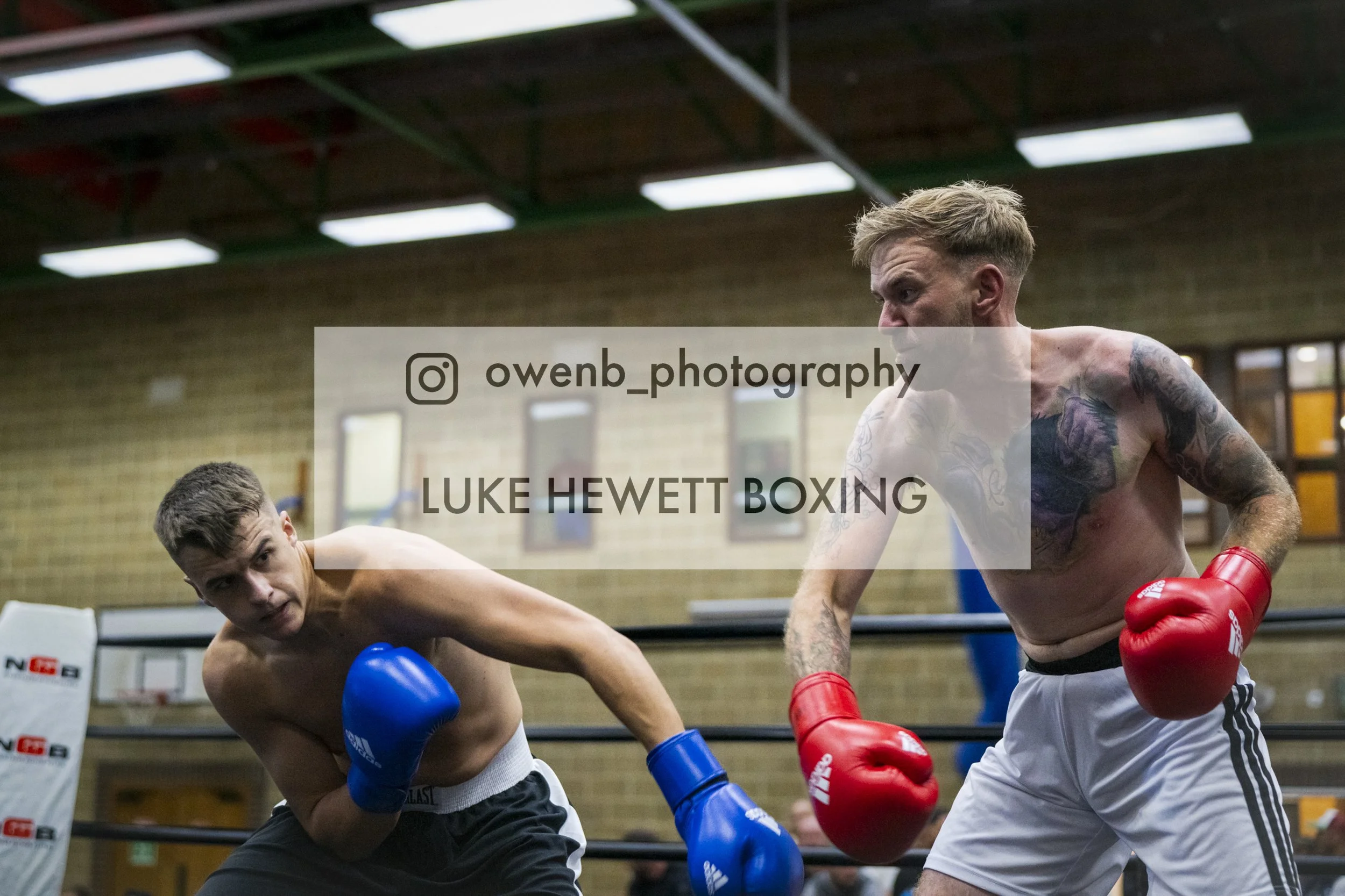 Two shirtless male boxers engaged in a match inside a boxing ring. One is leaning forward with blue gloves and the other with red gloves. They are indoors, in a gym with wooden walls and overhead lighting.