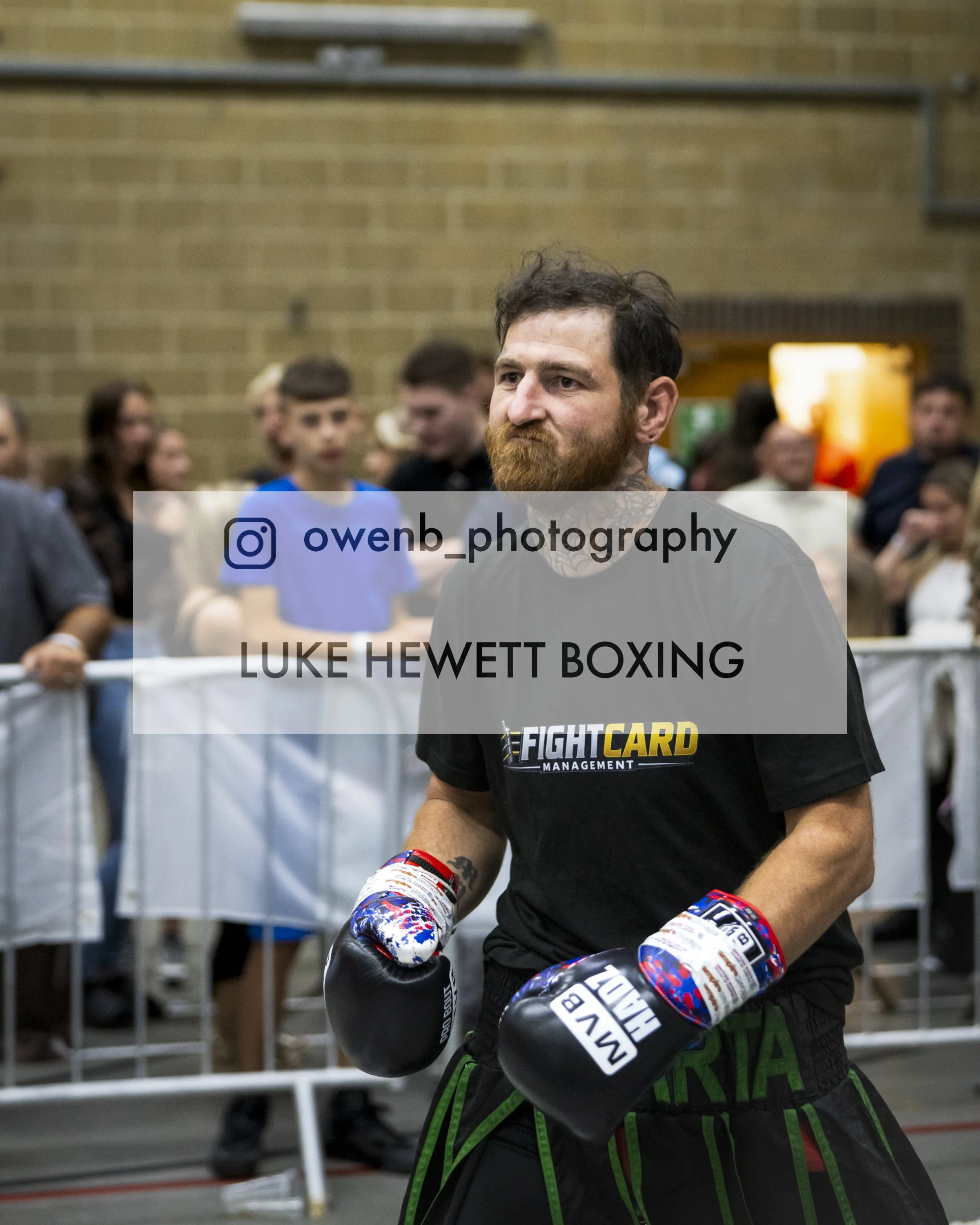 A male boxer wearing fight gloves stands in a boxing gym, with a serious expression, surrounded by people.