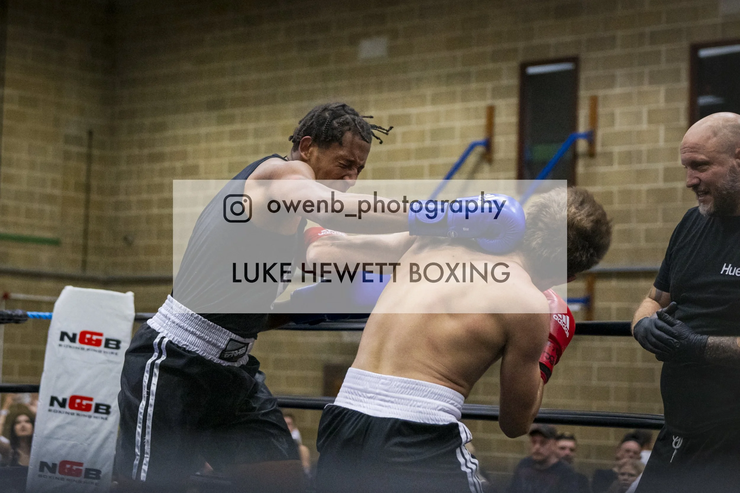 A boxing match where a boxer with a dark tank top and braided hair is punching another boxer with blonde hair, wearing red gloves, while an instructor or referee watches nearby. The scene takes place in a gym with brick walls.