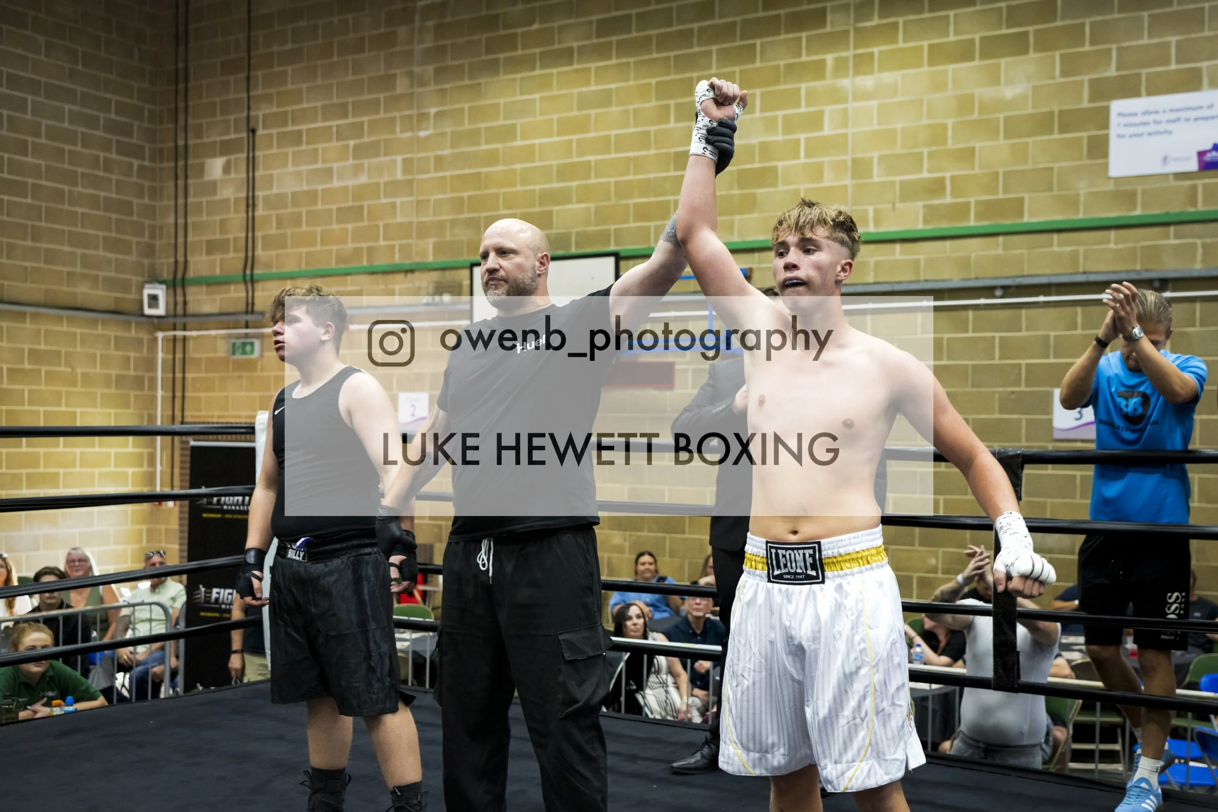 The photo shows a young male boxer with blond hair and light skin raising his arm in victory in a boxing ring, overseen by a man in black who is holding his hand up. The young boxer is shirtless, wearing white boxing shorts with yellow accents. In th
