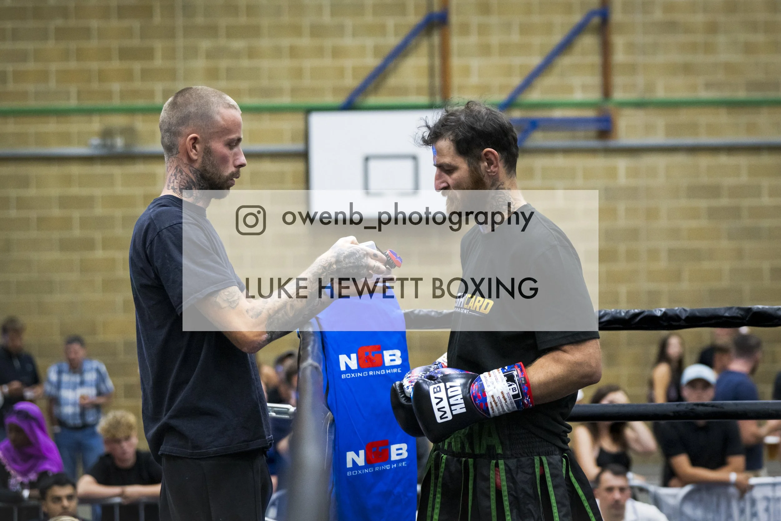 Two men in a boxing ring, one with a shaved head and tattoos, the other with dark curly hair, exchanging boxing gloves, with spectators watching in the background.