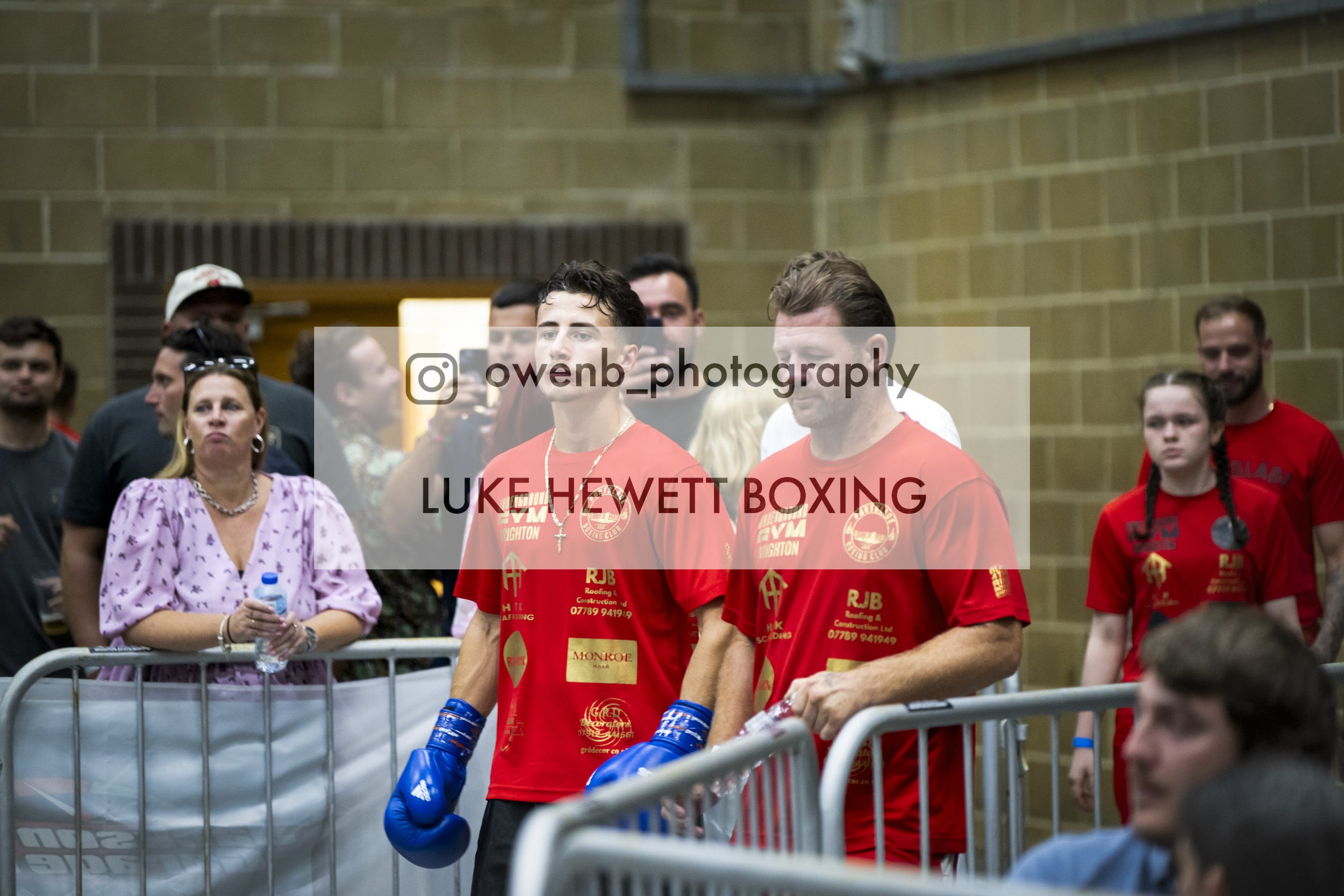 Group of people, including a woman in a purple dress holding a water bottle and a man in red boxing attire and blue gloves, standing behind a barrier in an indoor gymnasium with brick walls.