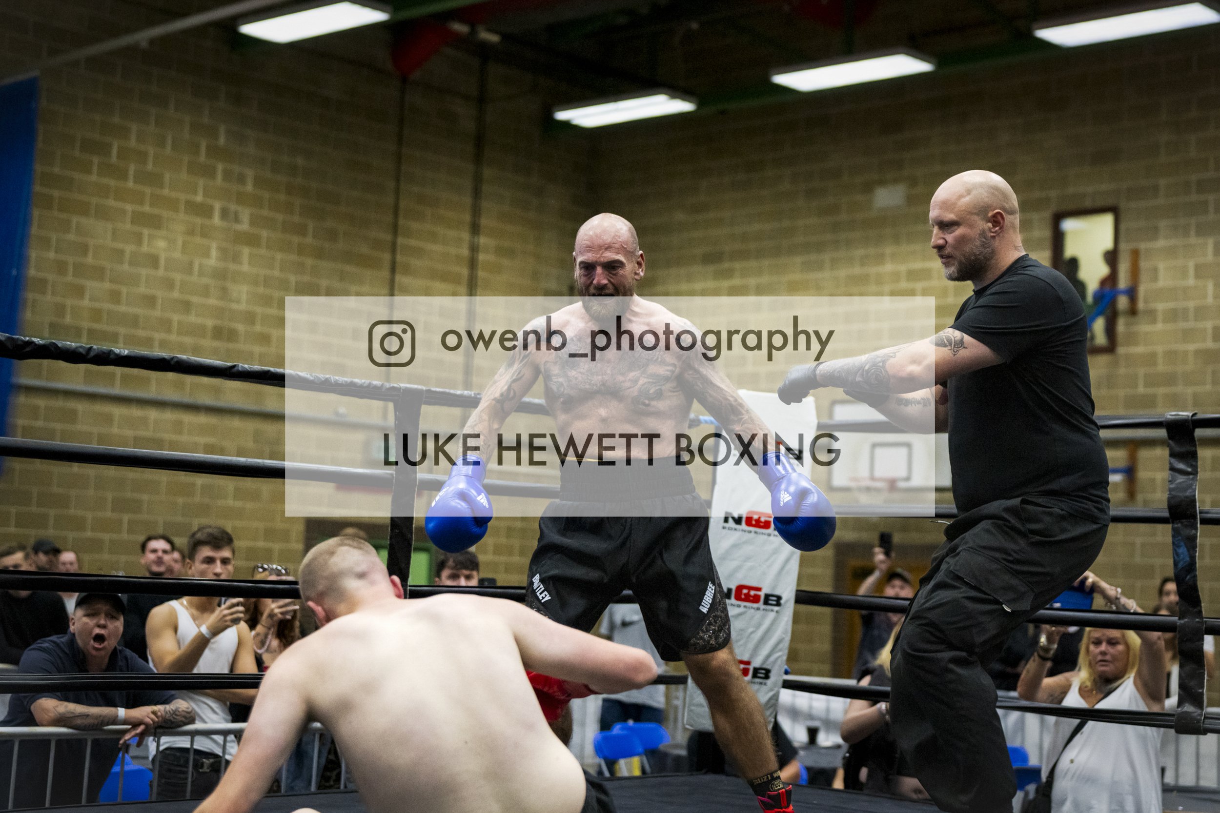 A boxing match taking place in a gym with a crowd watching. One fighter is shirtless with blue gloves, while the other is partially visible in the foreground. An adult male coach or trainer stands outside the ring, instructing the fighters.