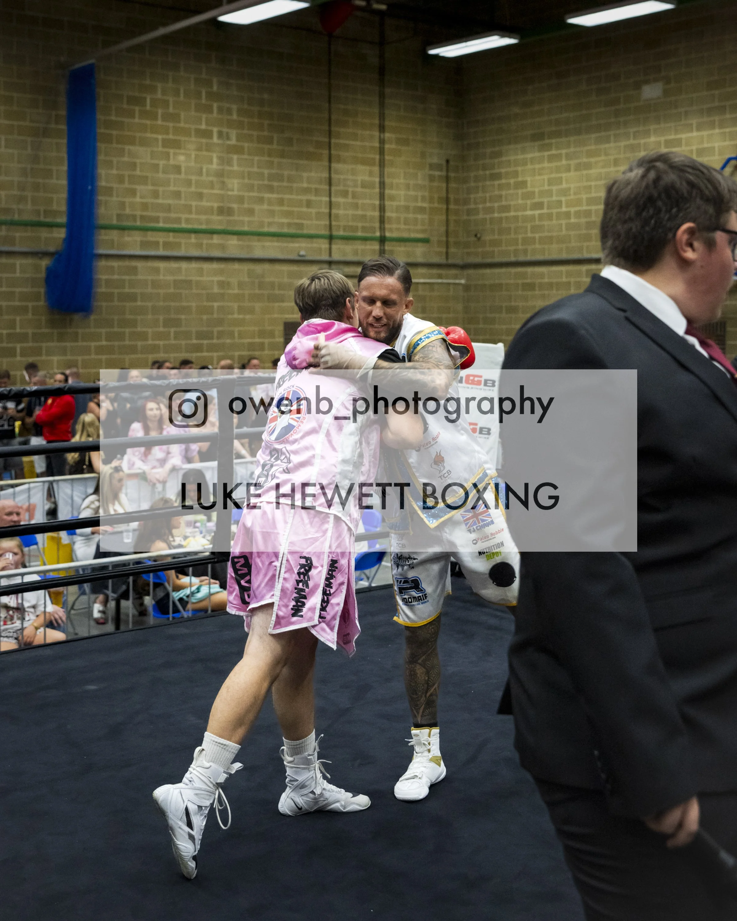 Two male boxers embracing after a match inside a boxing ring. One boxer is in a pink hoodie and shorts, the other is in a white and gold boxing outfit. There are spectators in the background and a person in a black suit in the foreground.