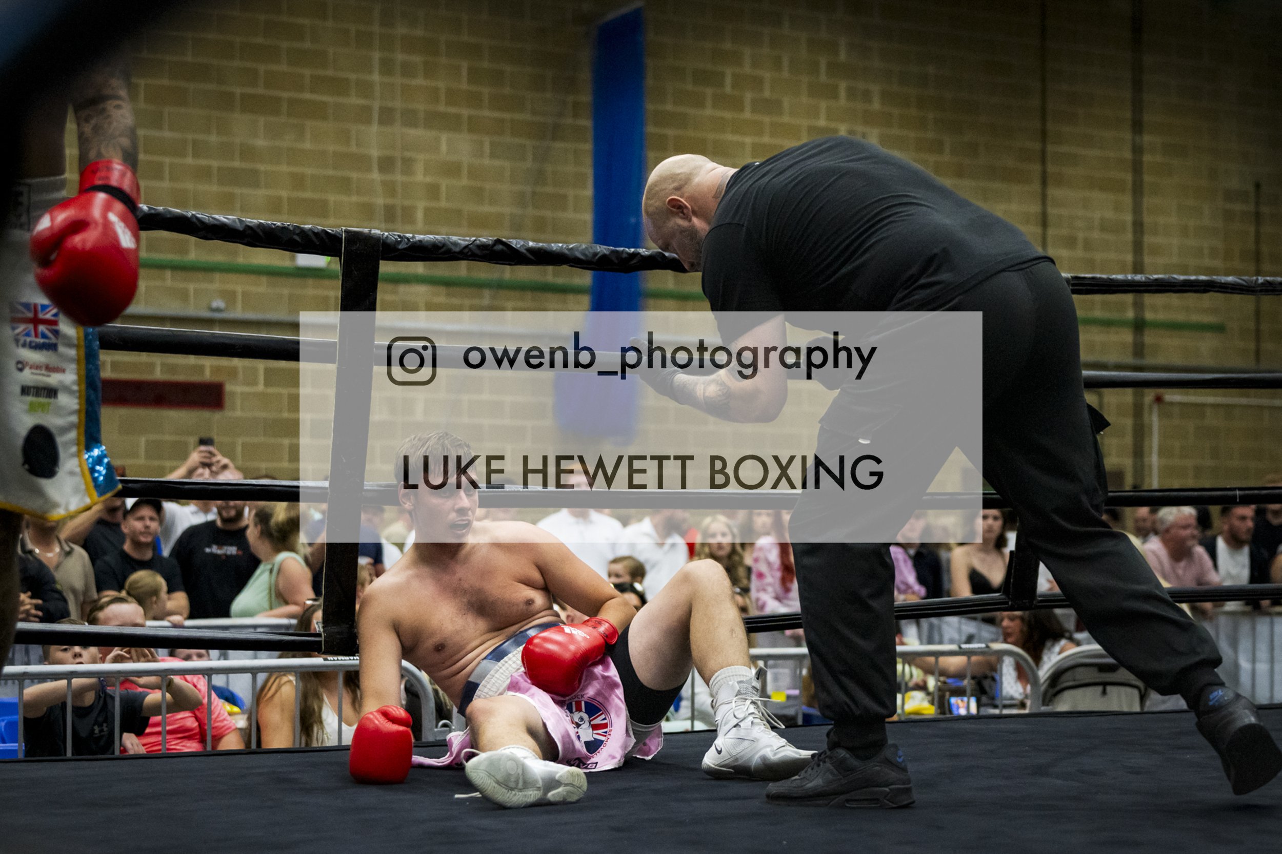 A boxer lies on the mat inside a boxing ring, looking up with his hand on his head, while a coach or referee leans over him. The boxer is wearing pink shorts, red gloves, and white shoes. There is a crowd gathered outside the ring, watching the scene
