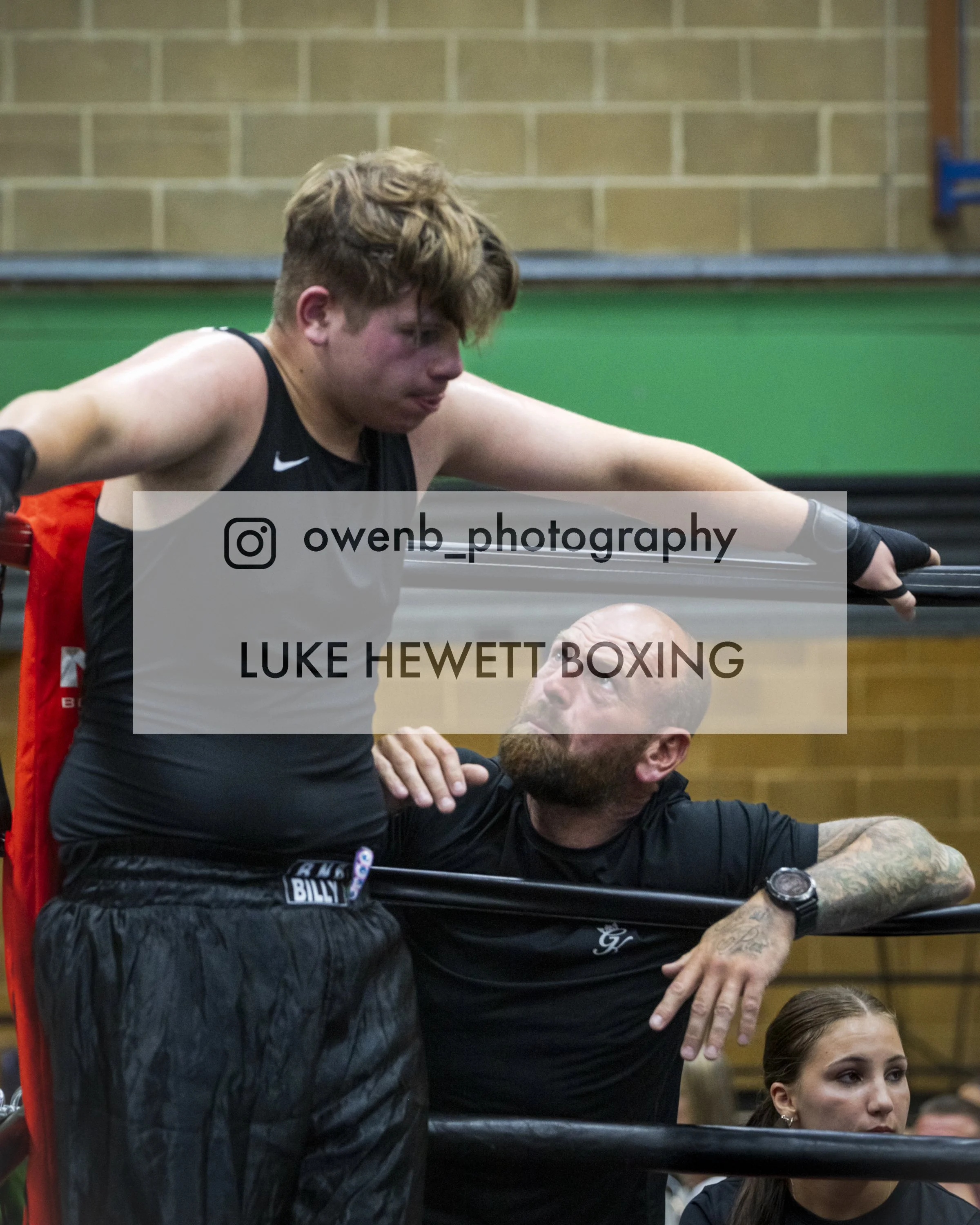 A young male boxer with light brown hair and tattoos on his arms, wearing a black shirt and black gloves, training with an older male coach in a boxing gym. The coach is wearing a black jacket and has tattoos on his forearms. They are practicing a bo