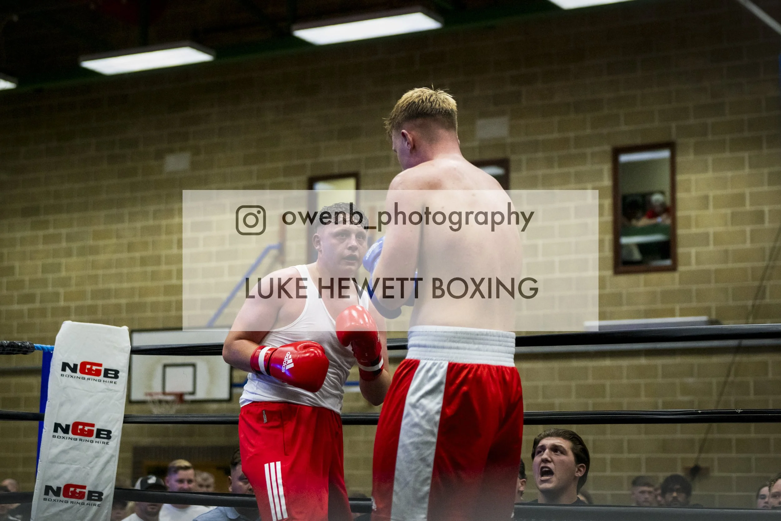 Two male boxers in red shorts and gloves sparring in a boxing ring, with an audience watching in the background and a brick wall behind them.