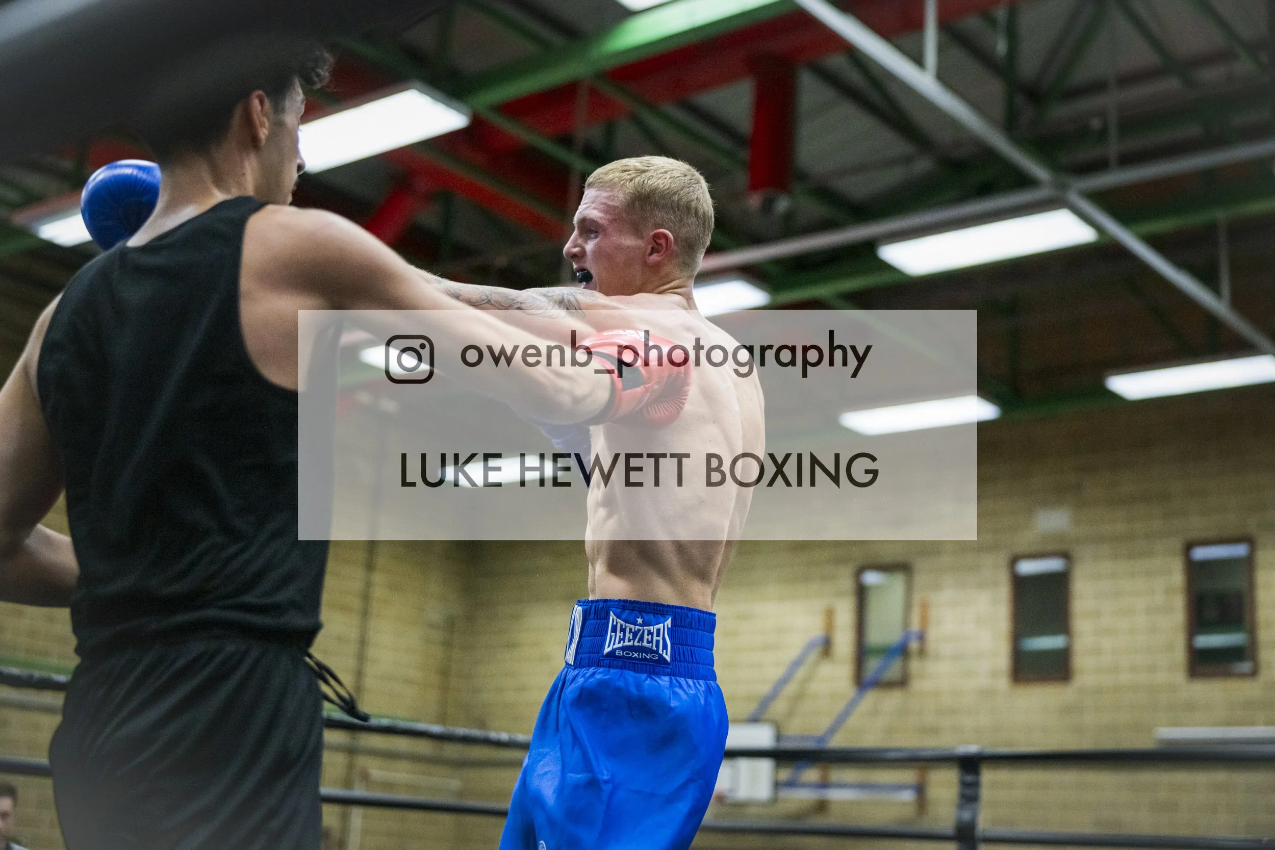 Two boxers training in a gym, one with a blue glove and the other with short blond hair, wearing blue shorts with 'GEZER BOXING' written on them, engaging in a sparring session.