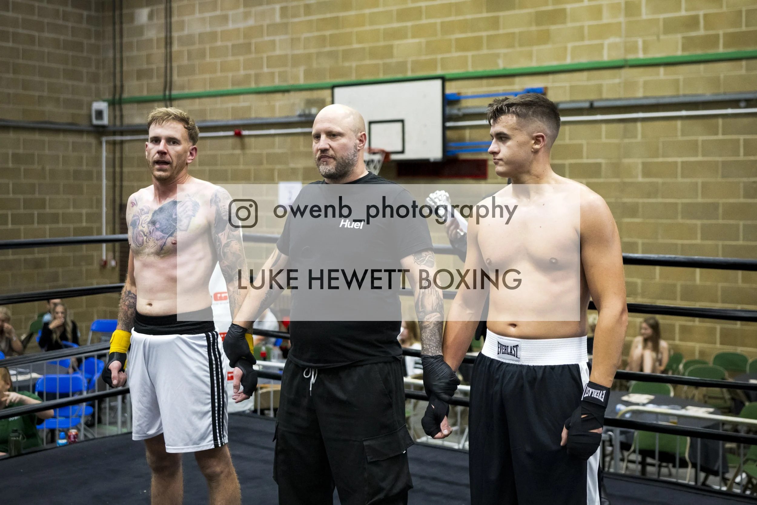 Two shirtless male boxers standing next to a referee in a boxing ring, with spectators seated behind the ring. The boxer on the left has tattoos and is wearing white shorts, while the boxer on the right has a fade haircut and is wearing black shorts.