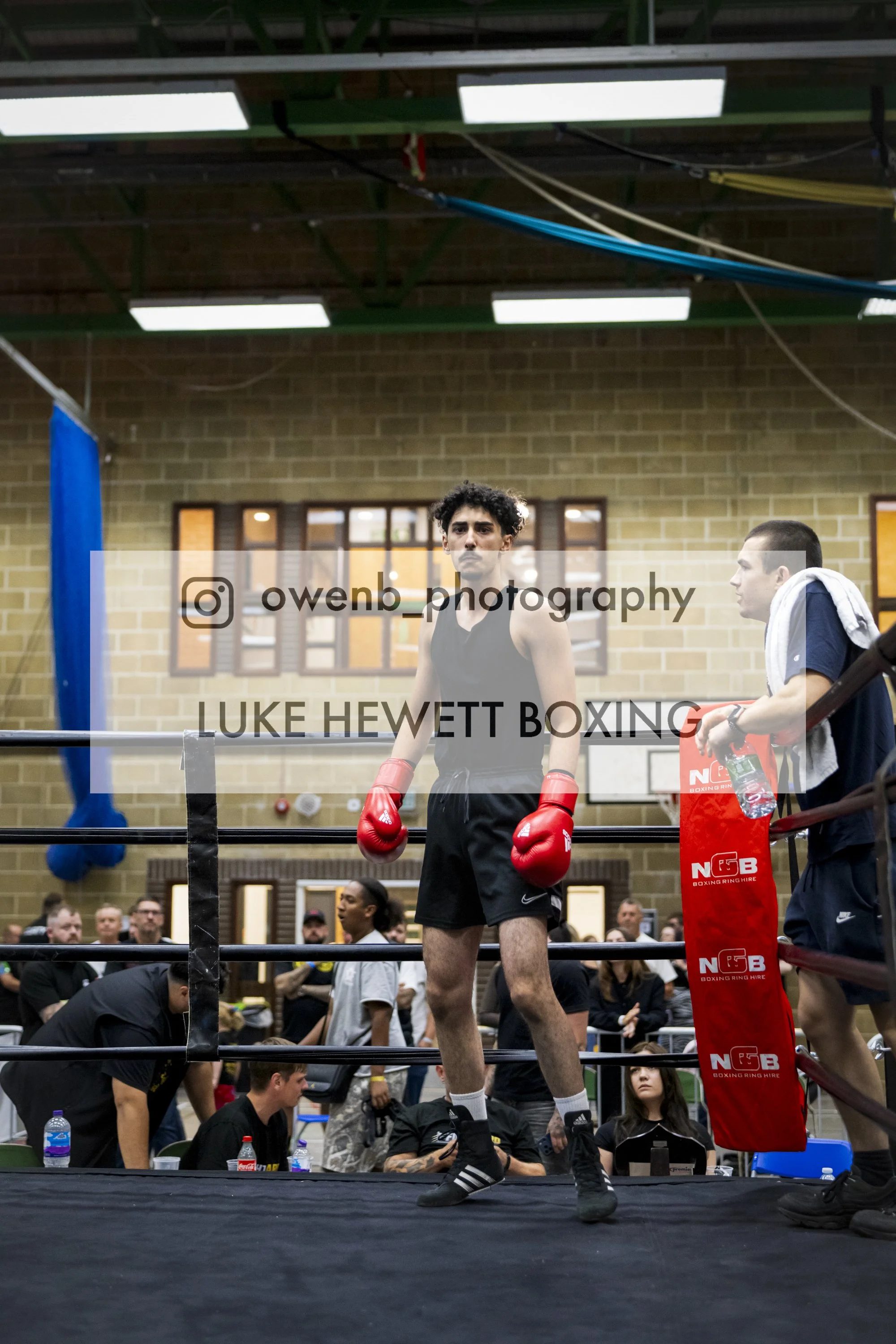 A young male boxer in black shorts and red gloves standing in the boxing ring, with an audience and staff visible behind him. The boxing ring has padded corner posts with red covers.