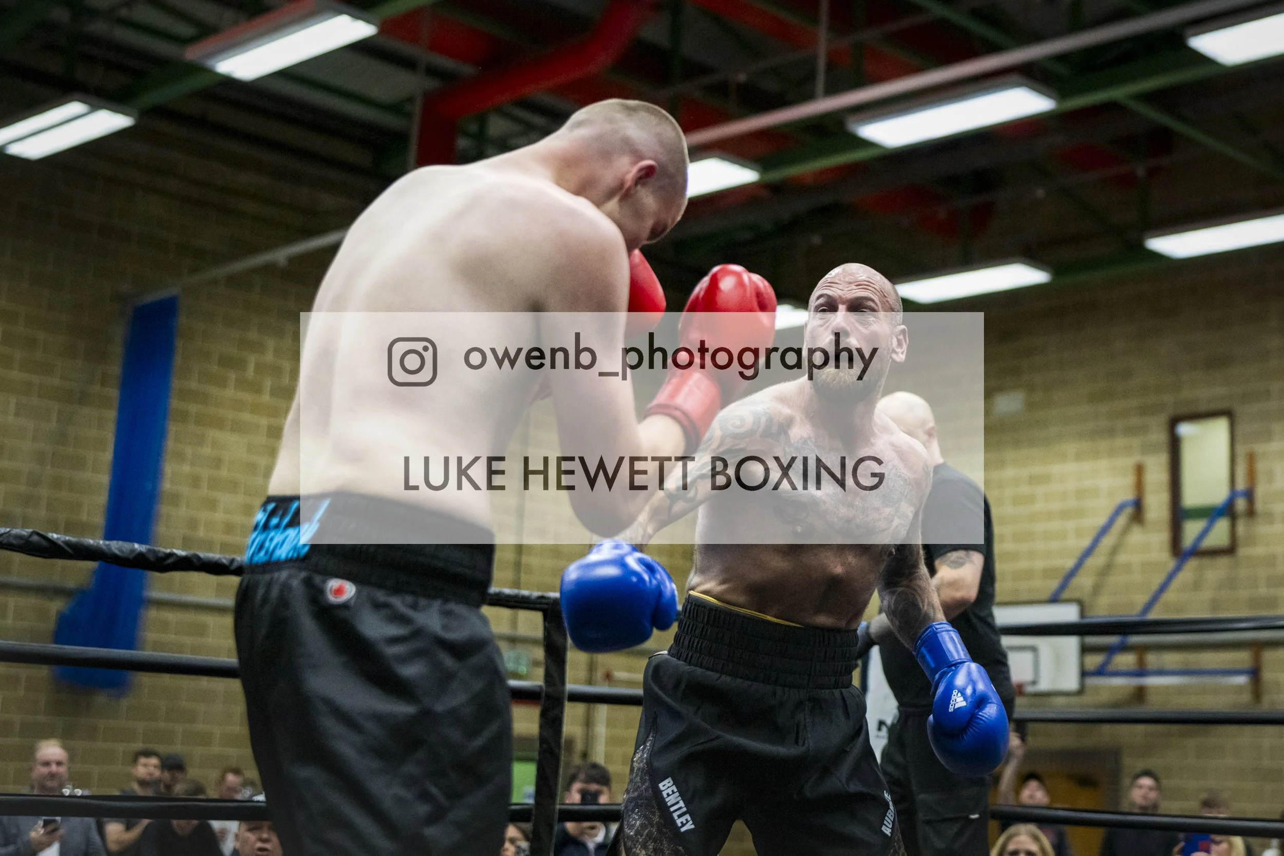 Two boxers sparring in a gym ring, one with red gloves and the other with blue gloves, with a crowd watching in the background.