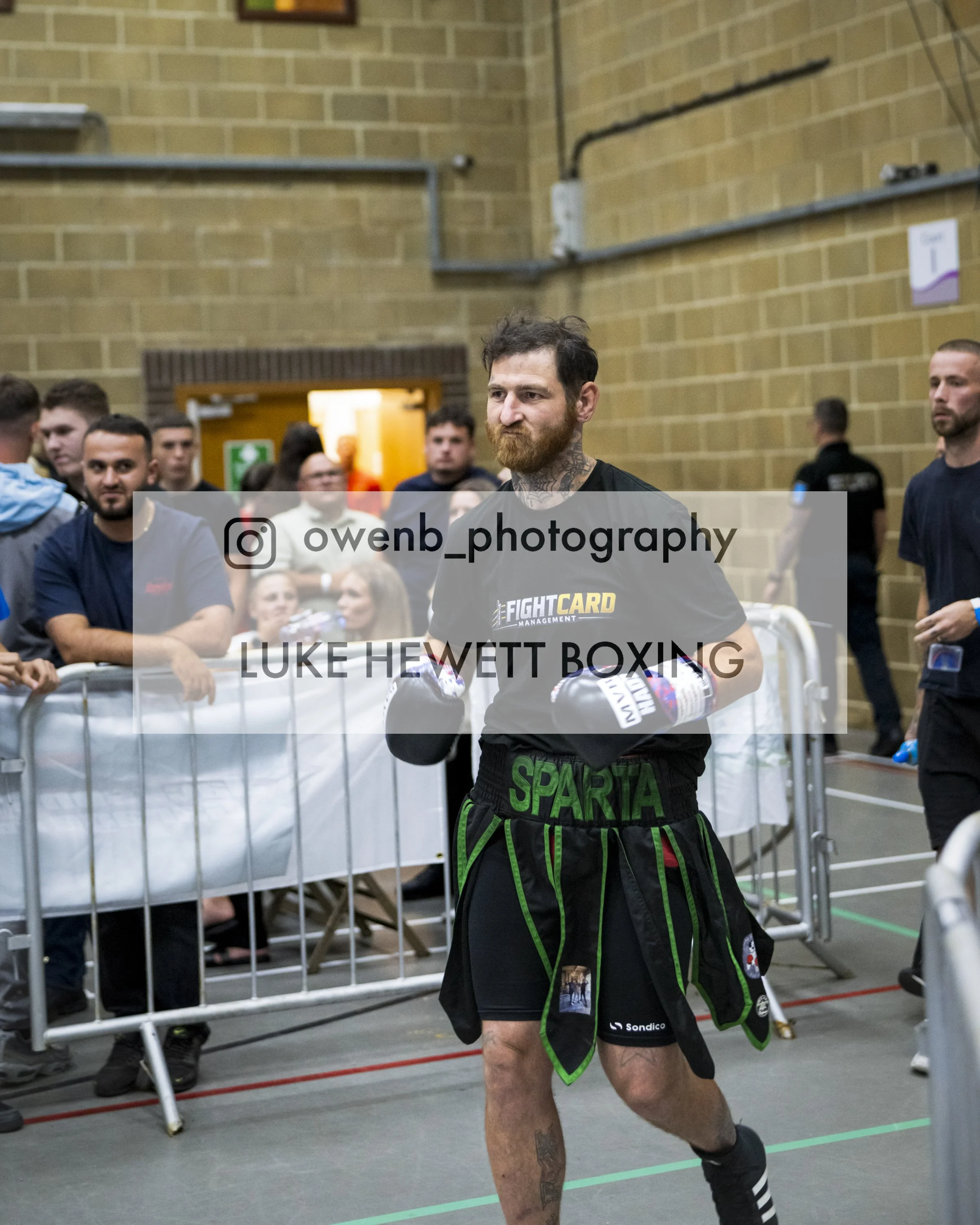 A male boxer with a beard and tattoos on his neck and legs, wearing boxing gloves and shorts, is walking into a boxing ring area with a crowd of spectators and other individuals in the background. The venue has a brick wall and industrial pipes. The 