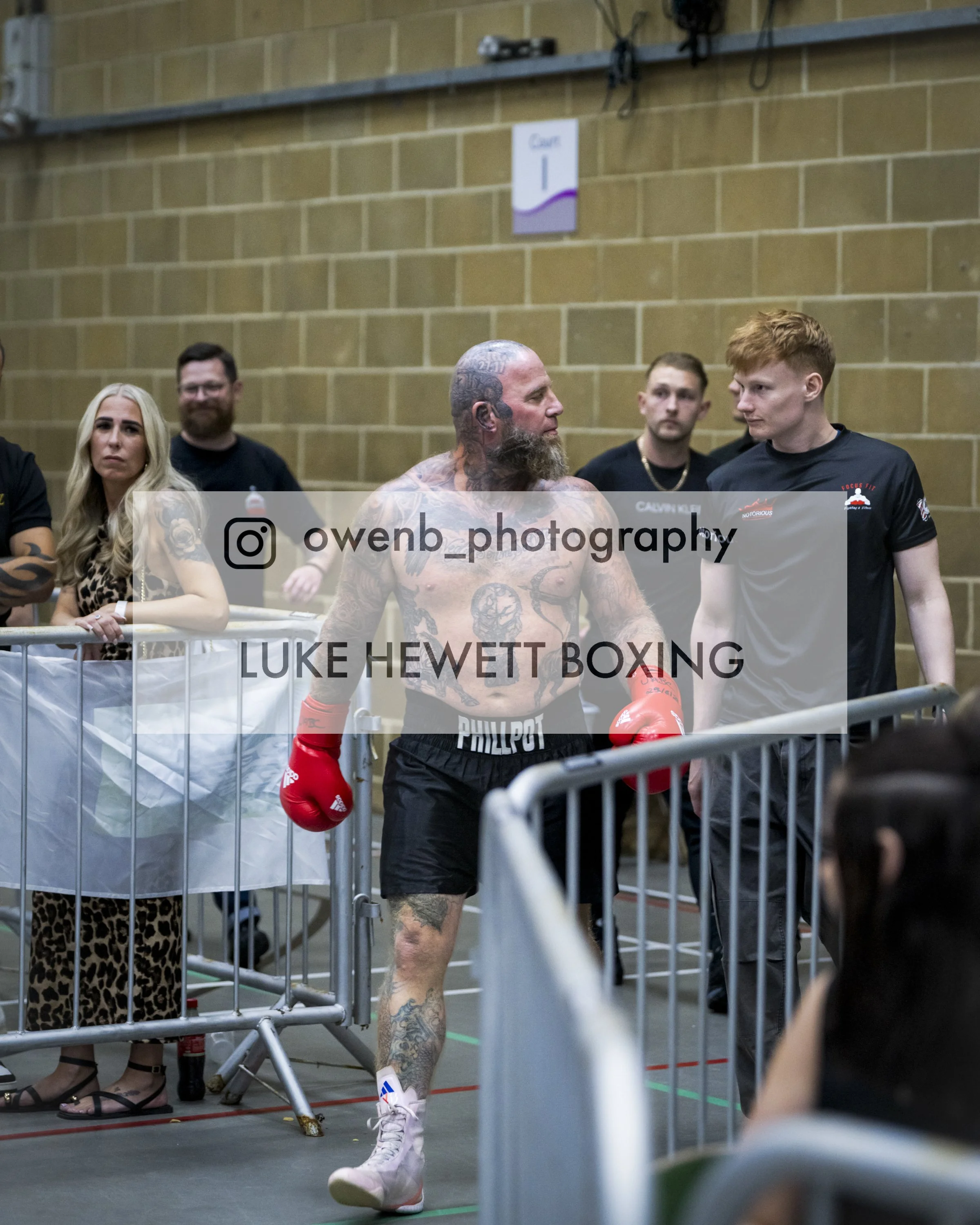 Boxer with tattoos, wearing red gloves, white shoes, and black shorts, walking in the ring backstage at a boxing event, with people and a cardboard barrier in the background.