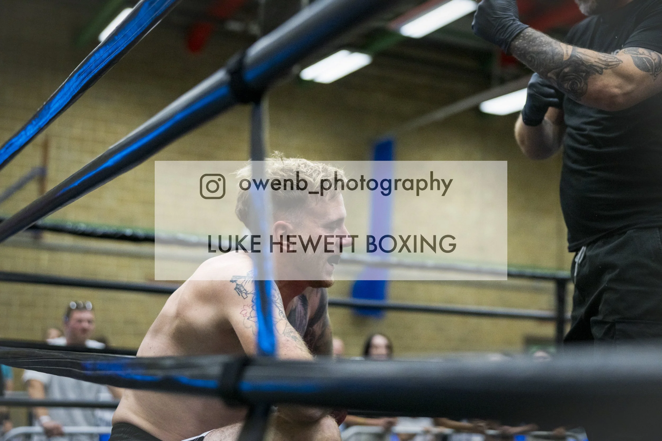 Boxer Luke Hewett resting in a boxing ring during training, with a trainer and spectators in the background.