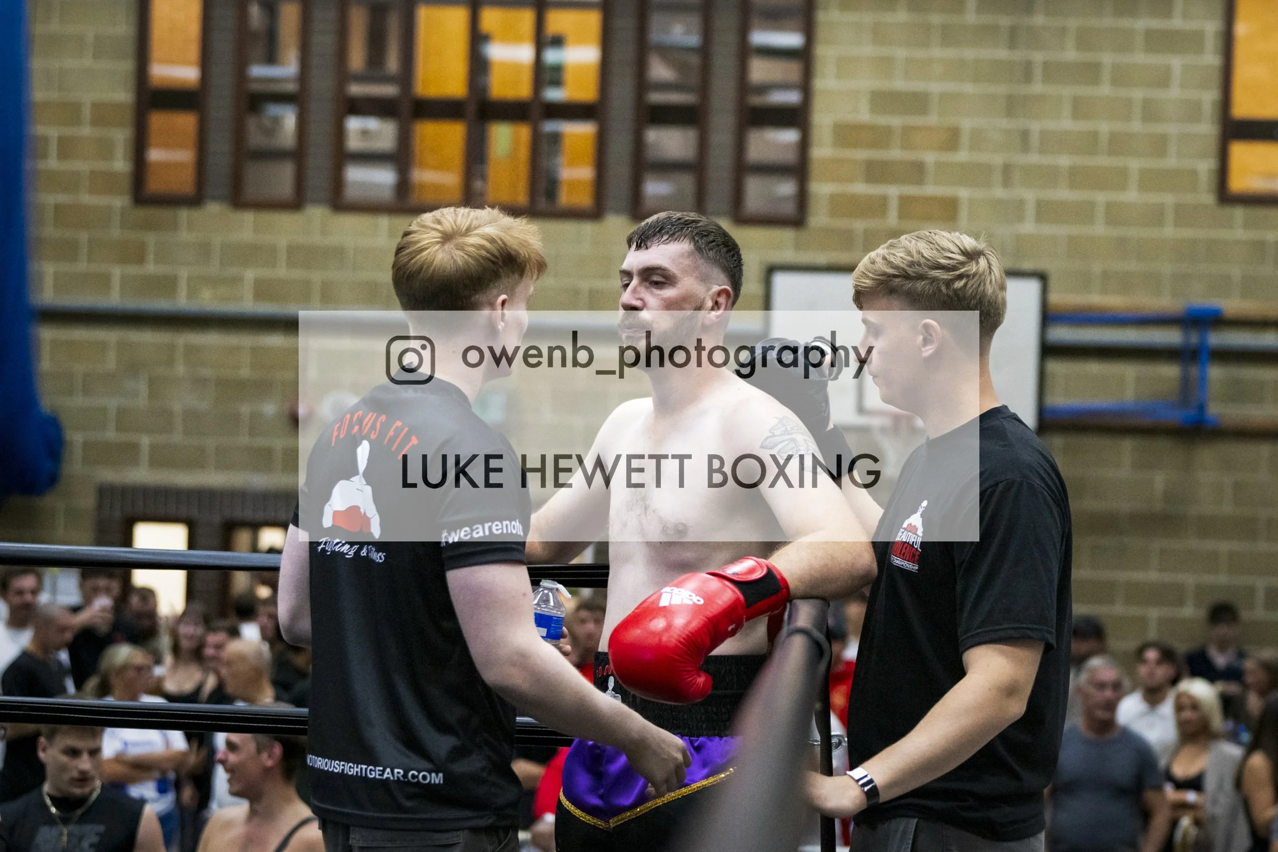 A boxer wearing red gloves and black shorts is in a boxing ring, talking with a trainer who is shirtless and holding a water bottle. Another person with short blonde hair is standing nearby. The background shows an audience in a gymnasium with brick 