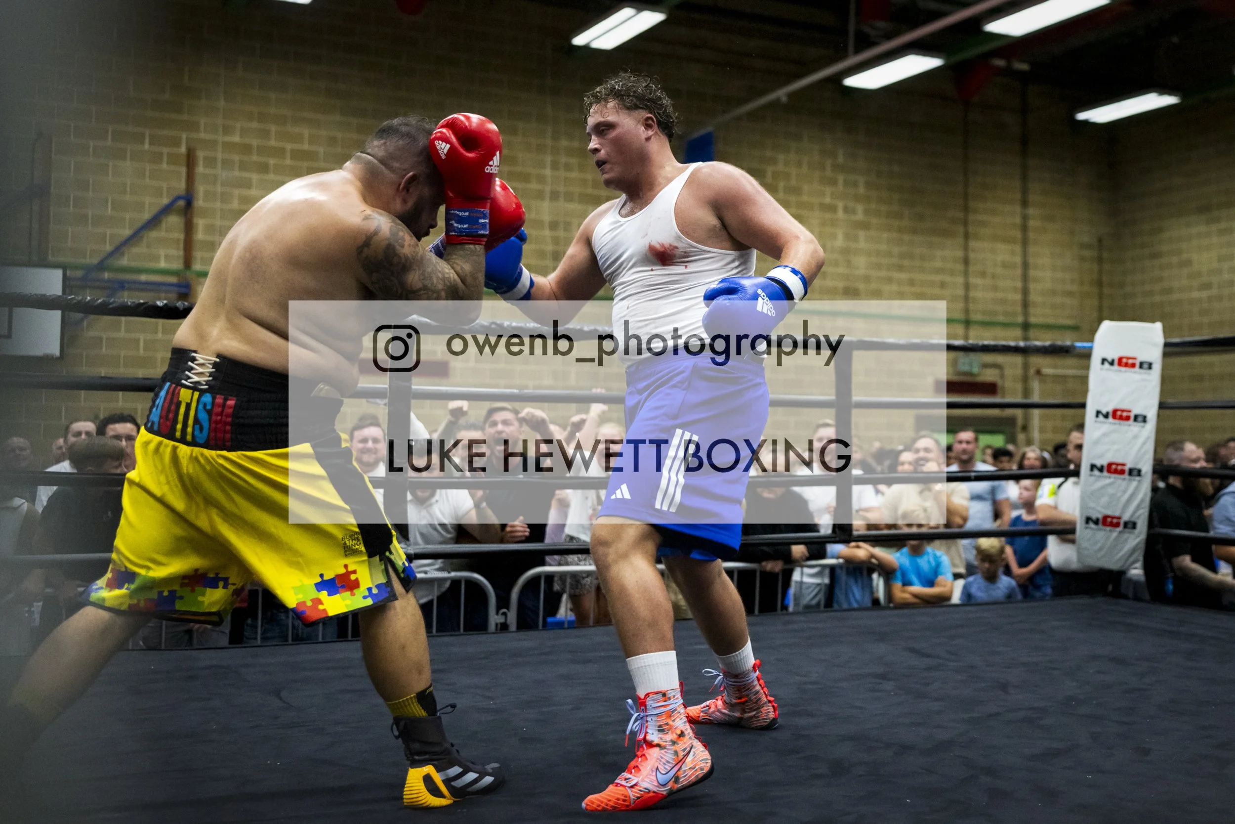 Two men boxing in a ring, one in yellow shorts and the other on the ropes, both wearing boxing gloves, with spectators watching in the background.