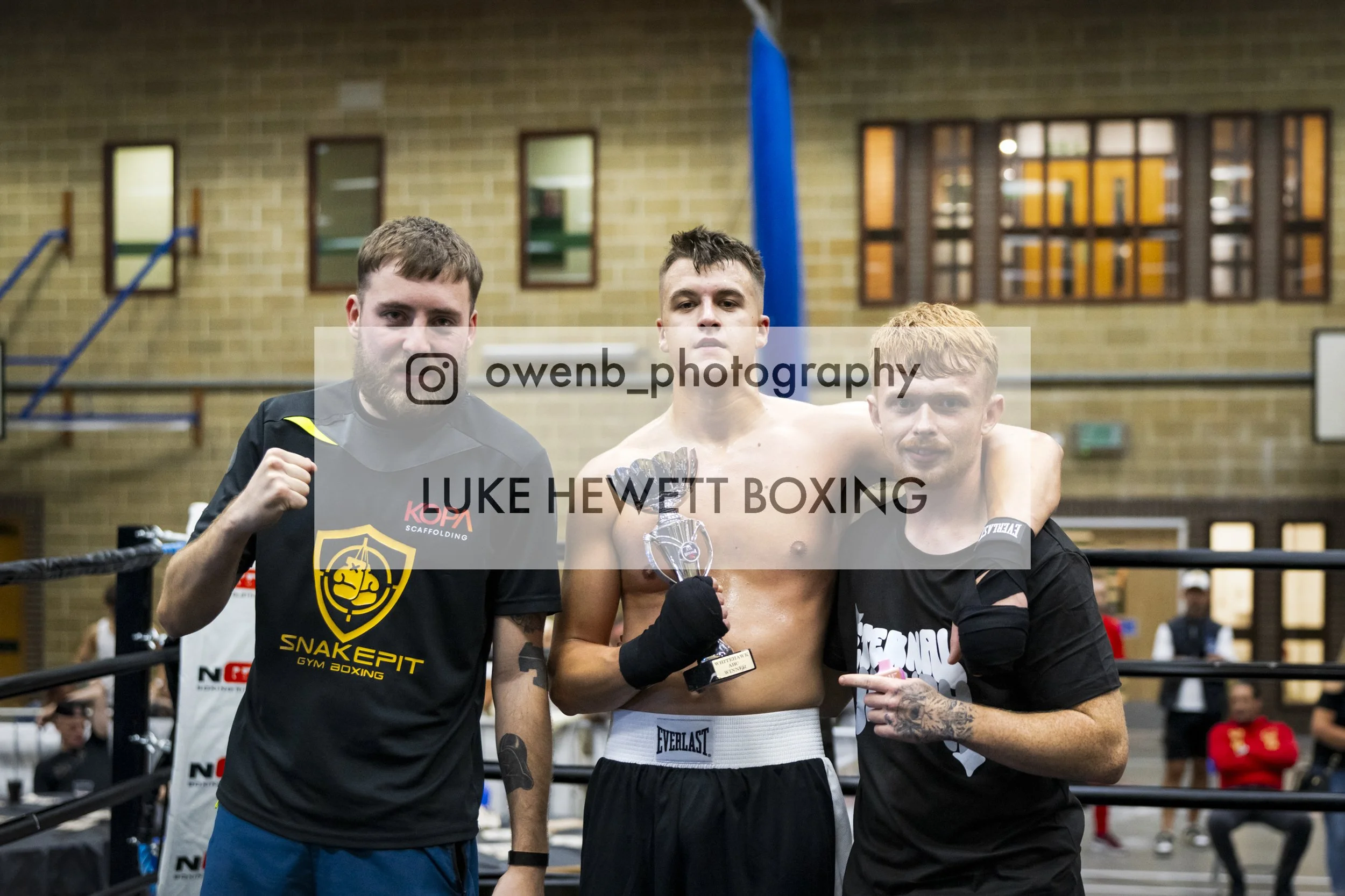 Three male boxers standing in the boxing ring, with the boxer in the middle holding a trophy, the other two men are posing with him. The boxers are wearing gloves and athletic clothing. The background shows gym surroundings with windows and spectator
